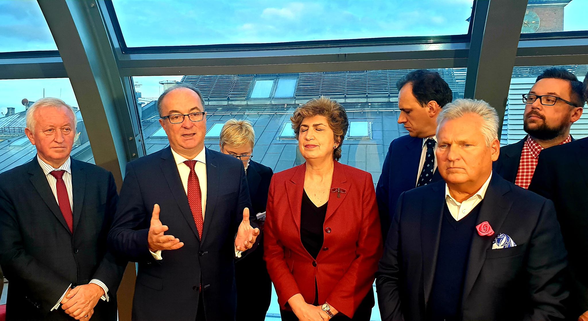 Grupa polityków w garniturach stoi w nowoczesnym, przeszklonym wnętrzu podczas konferencji; mężczyzna po lewej gestykuluje, a kobieta w czerwonej marynarce stoi obok.