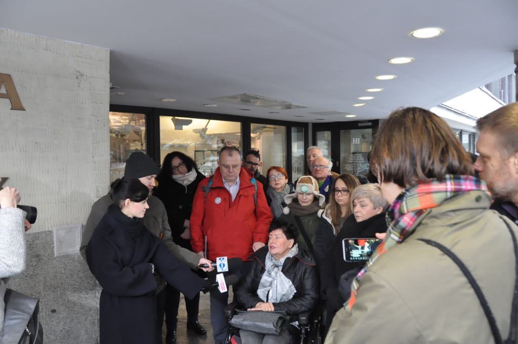 Grupa ludzi przed wejściem do budynku, w centrum osoba na wózku rozmawia z dziennikarzami i kamerzystami podczas inauguracji obywatelskiej inicjatywy ustawodawczej dotyczącej świeckiego państwa.
