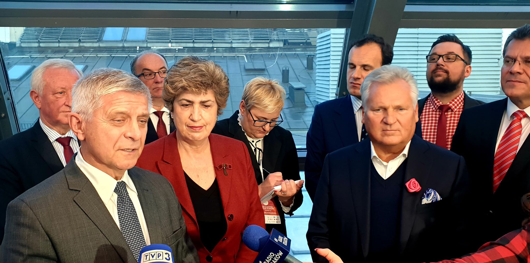 Grupa polityków w garniturach udziela konferencji prasowej, stojąc przed mikrofonami dziennikarzy w jasnym, nowoczesnym wnętrzu.