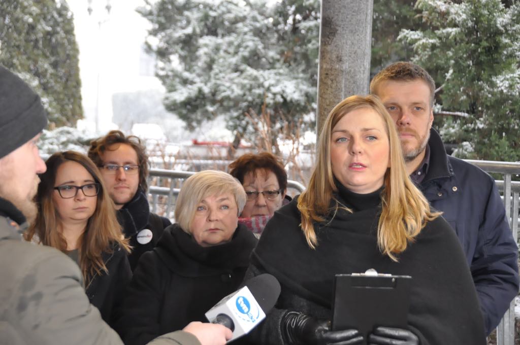Kobieta z clipboardem przemawia do mikrofonu podczas konferencji prasowej o świeckim państwie, otoczona innymi uczestnikami w zimowej scenerii.