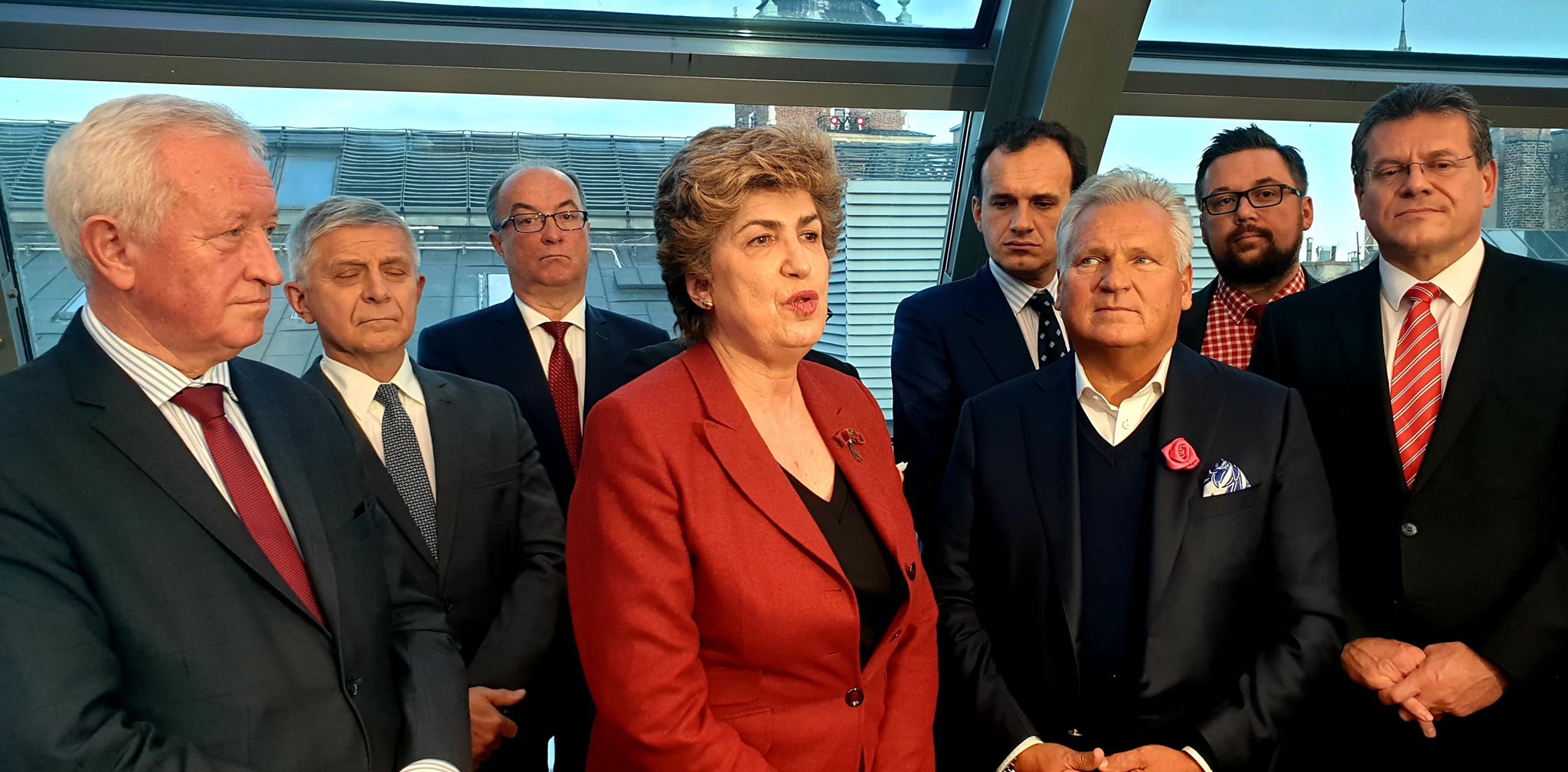 Grupa polityków w garniturach, z kobietą w czerwonym żakiecie na pierwszym planie, przemawia podczas spotkania w wnętrzu z przeszklonym sufitem.