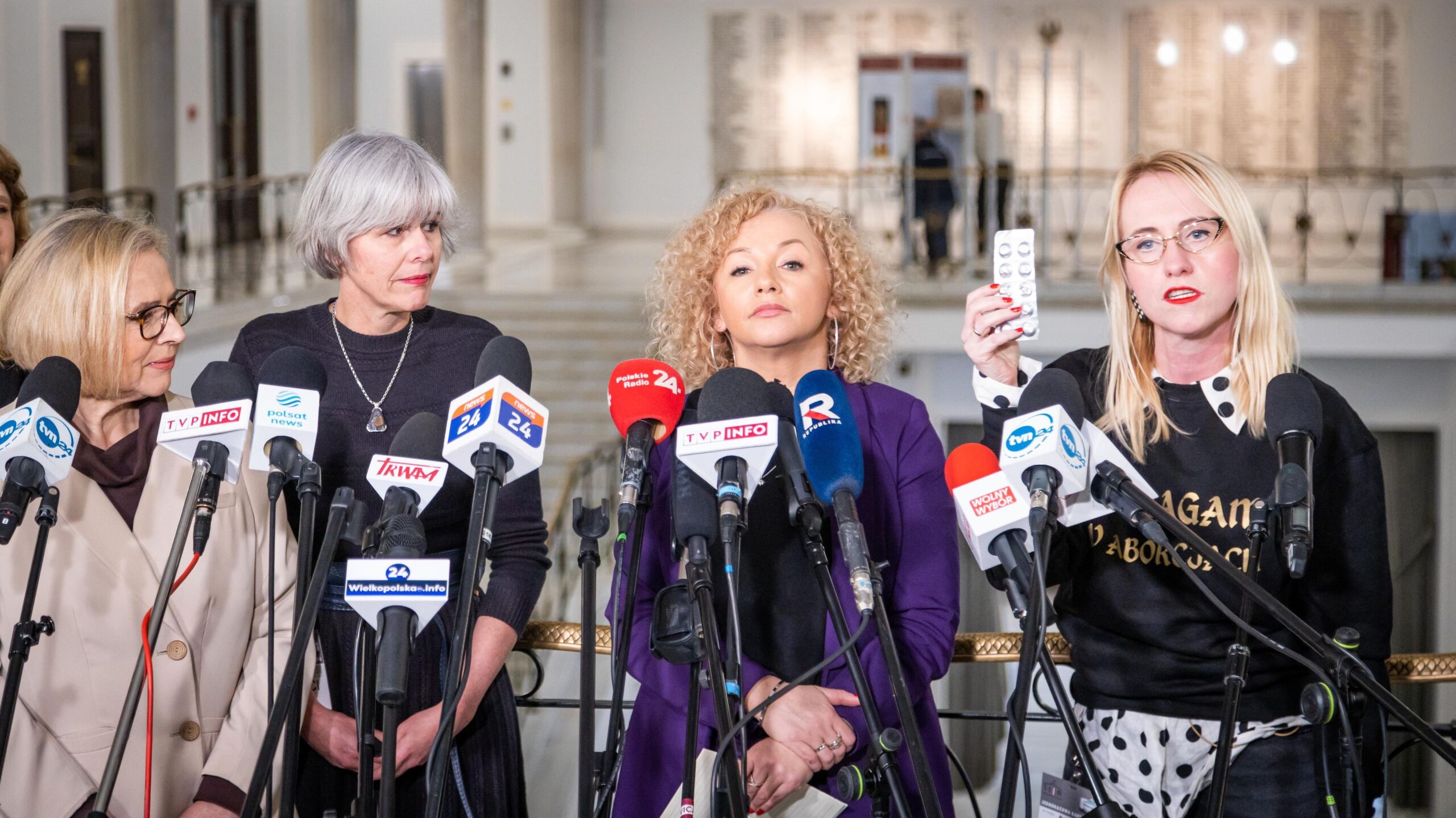 Cztery kobiety stoją na konferencji prasowej, otoczone licznymi mikrofonami; po prawej kobieta trzyma w dłoni blister z tabletkami.
