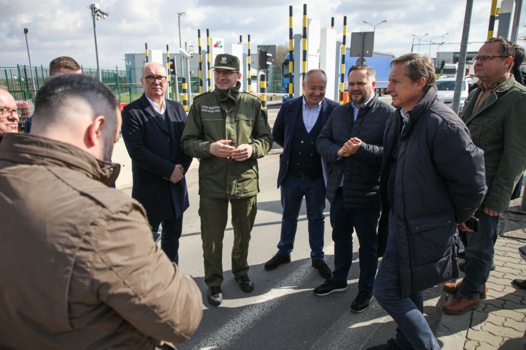 Na zdjęciu widoczna jest grupa mężczyzn, w tym jeden w mundurze, rozmawiających w otoczeniu infrastruktury, w słoneczny dzień.