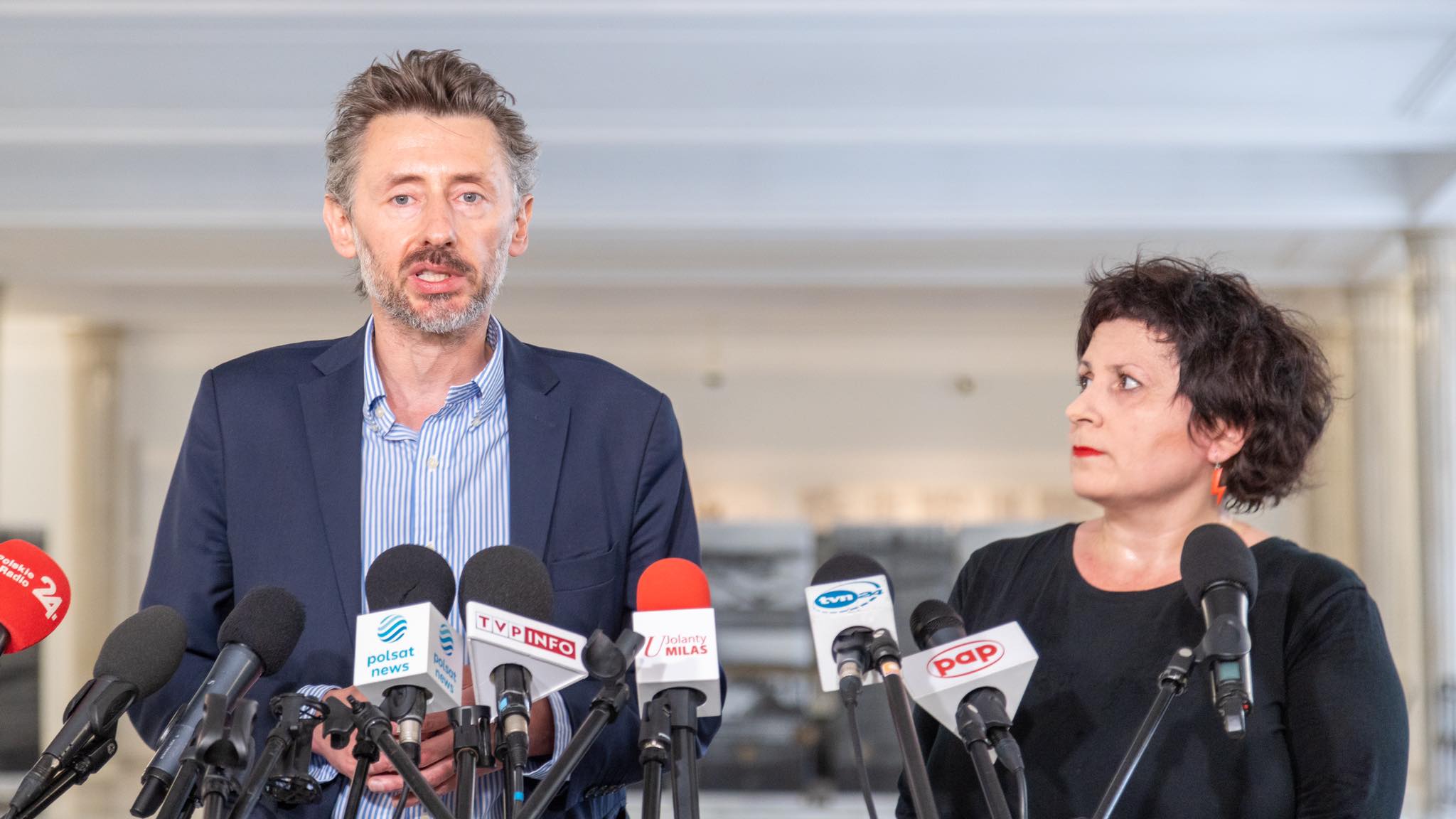 Na zdjęciu widoczna jest osoba przemawiająca przy mównicy otoczonej mikrofonami, a obok niej stoi druga osoba słuchająca z uwagą.