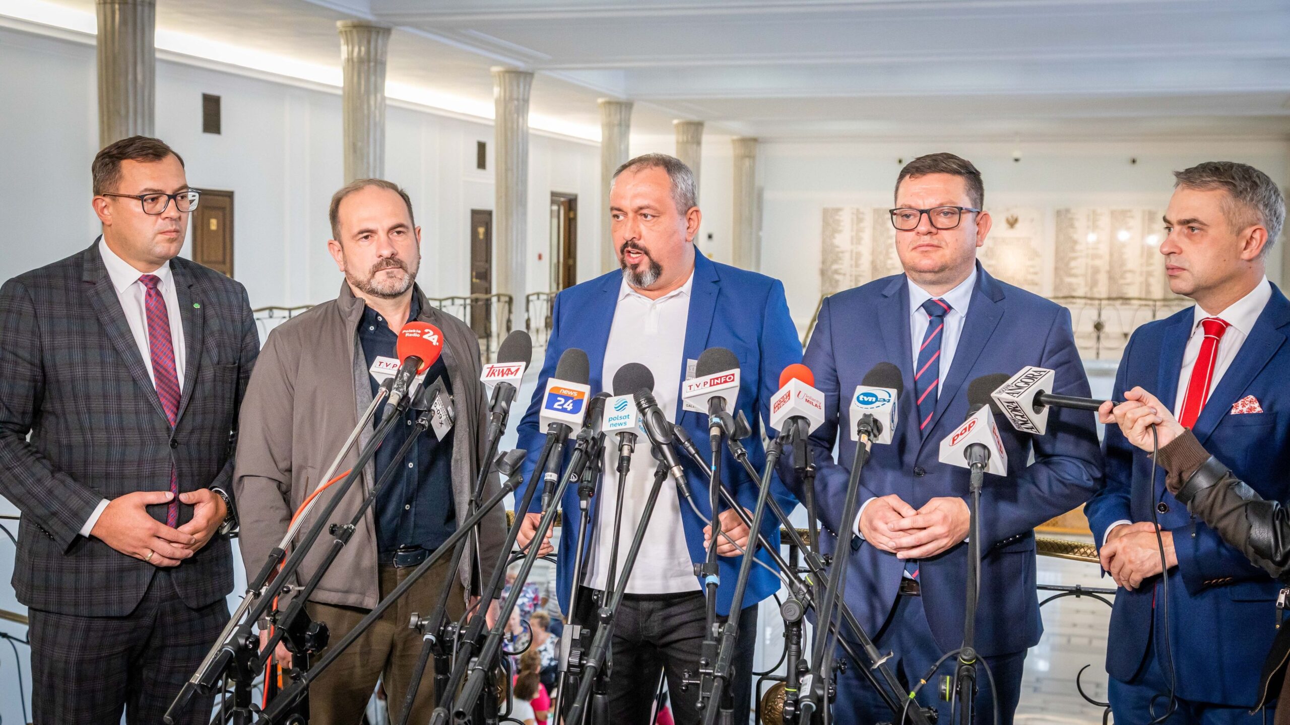 Konferencja prasowa z udziałem kilku mężczyzn stojących przy mikrofonach w gmachu instytucji publicznej.