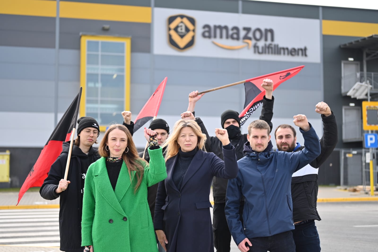 Grupa protestujących stoi przed budynkiem Amazon Fulfillment w Polsce, trzyma flagi i podnosi pięści, domagając się kontroli PIP we wszystkich magazynach Amazon Polska.