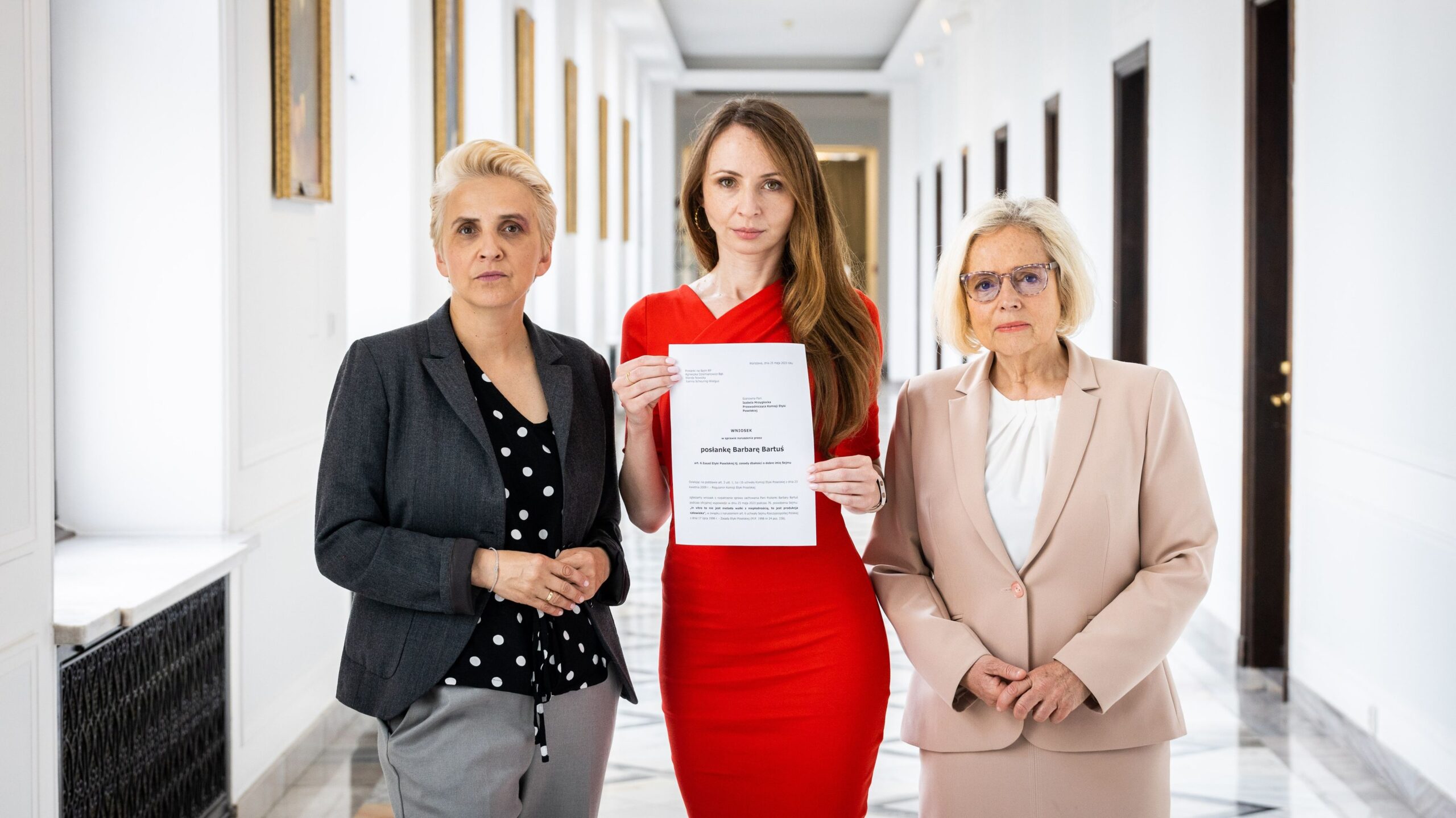 Trzy kobiety w eleganckich strojach stoją w jasnym korytarzu; środkowa kobieta w czerwonej sukience trzyma kartkę z dokumentem.