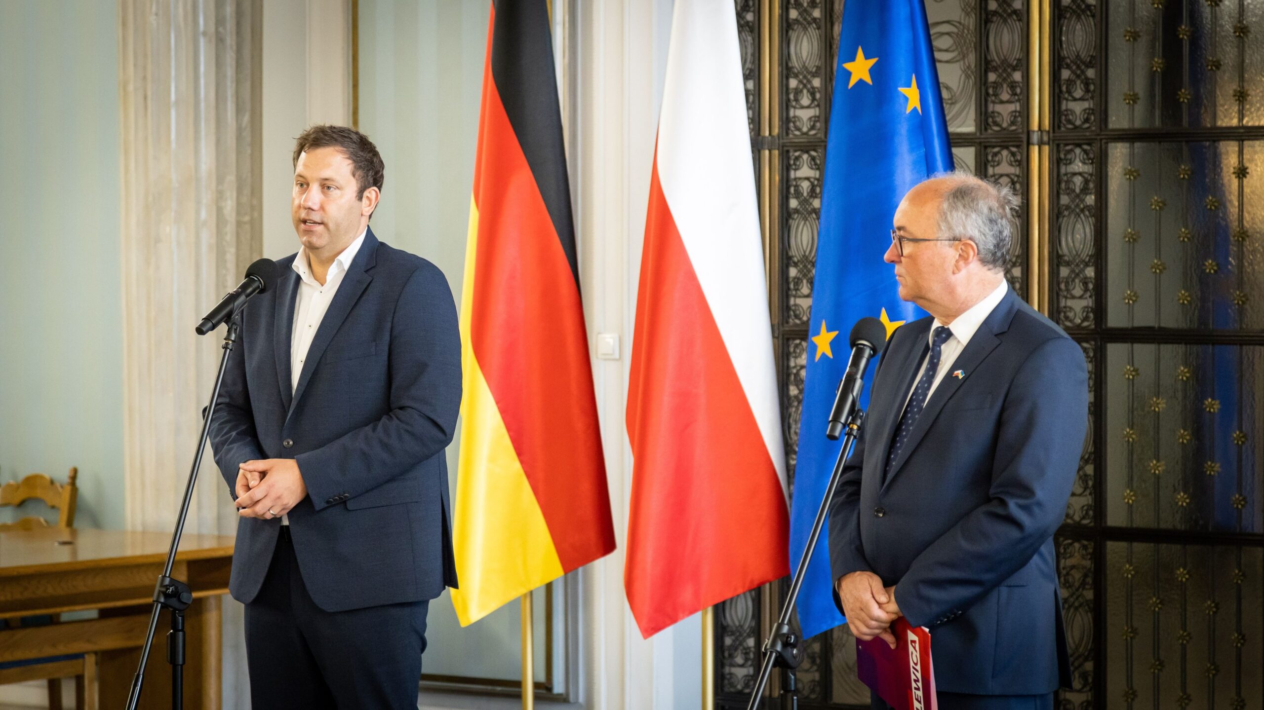 Dwóch mężczyzn w garniturach przemawia do mikrofonów podczas konferencji, stojąc obok flag Niemiec, Polski i Unii Europejskiej.