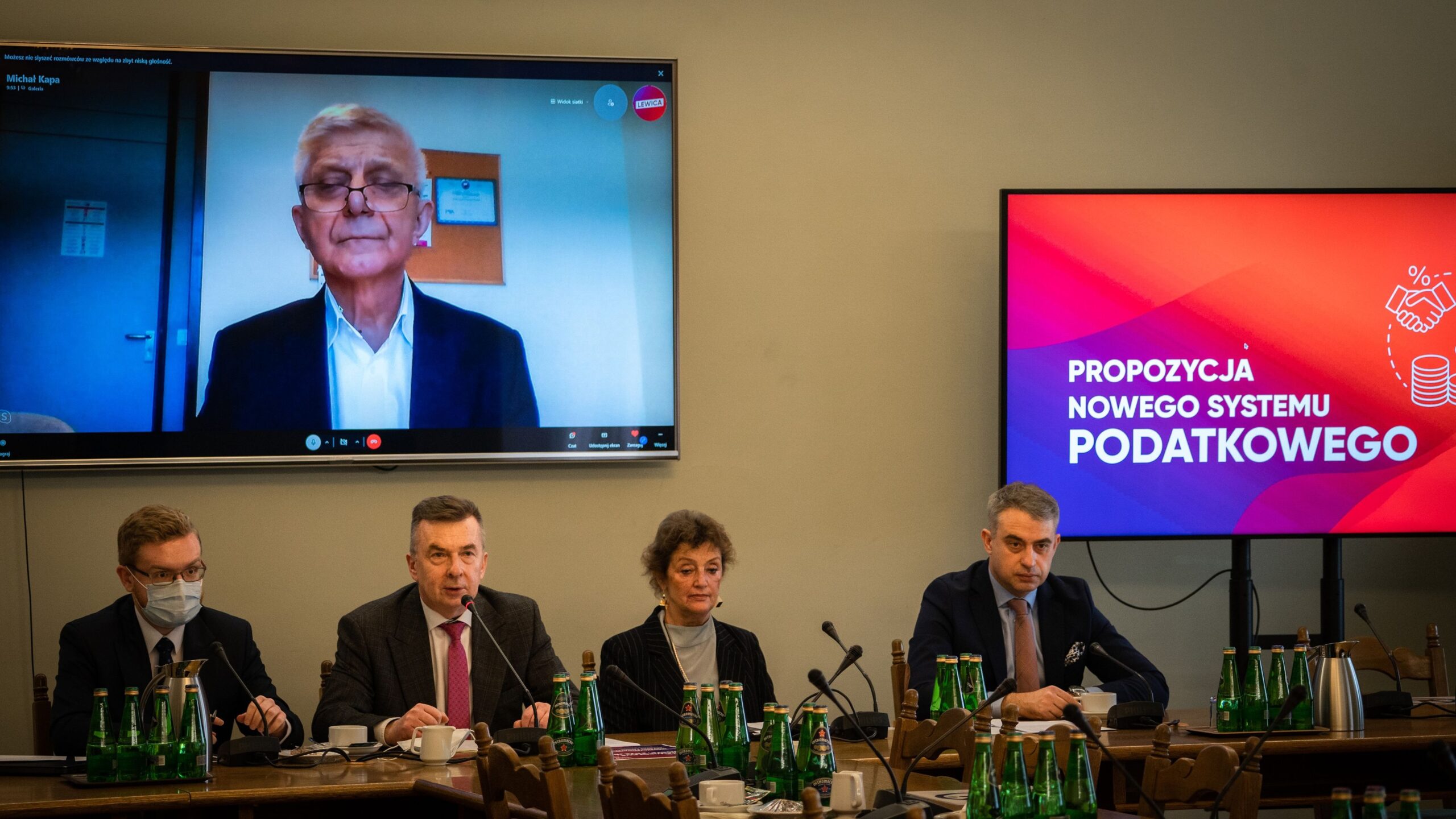 Na zdjęciu przedstawiono spotkanie dotyczące propozycji nowego systemu podatkowego, z uczestnikami w sali i jednym mówcą obecnym przez wideo.