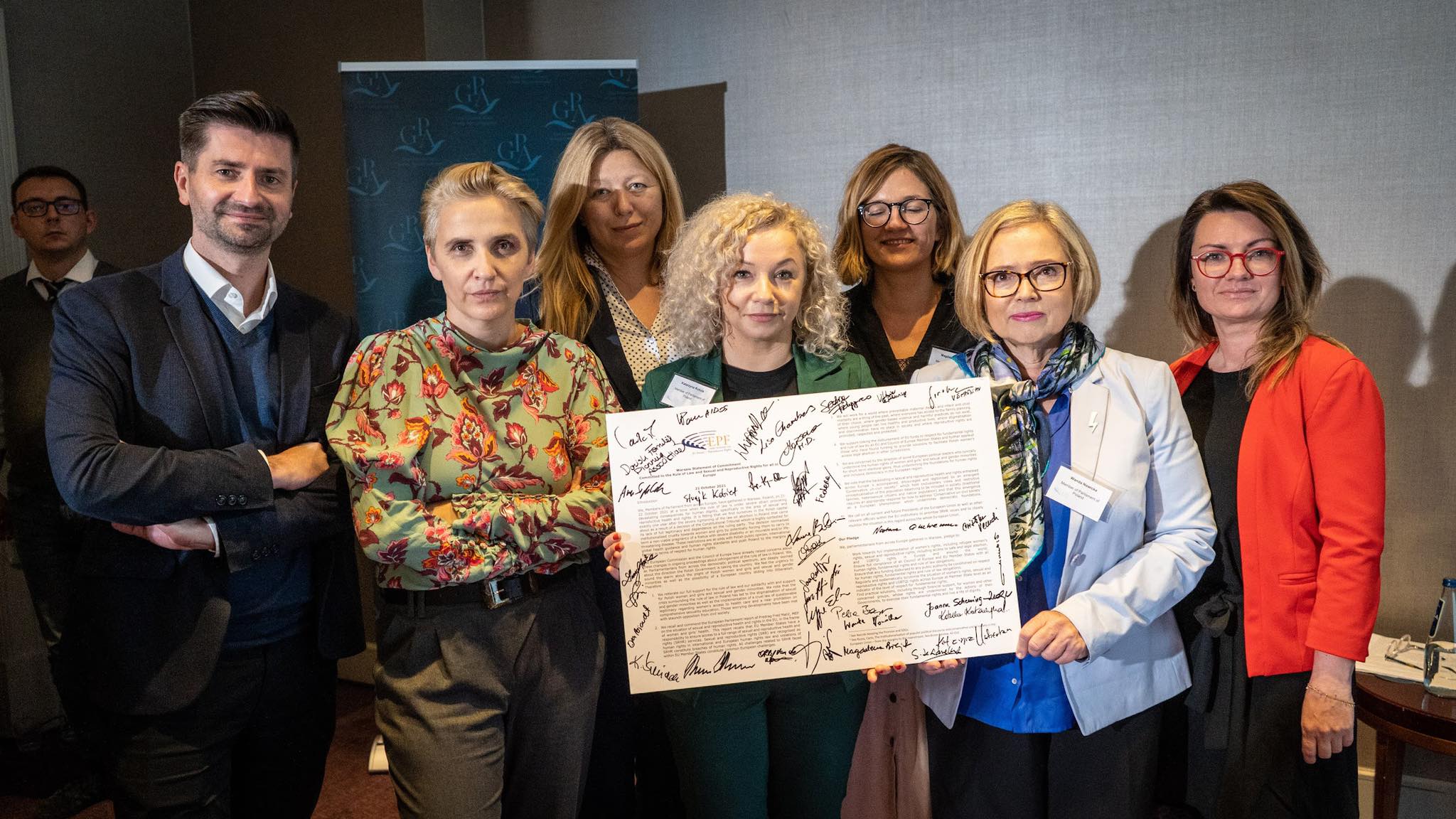 Grupa siedmiu osób na konferencji stoi razem i prezentuje dużą kartę z podpisami, stojąc na tle banera z logotypem.