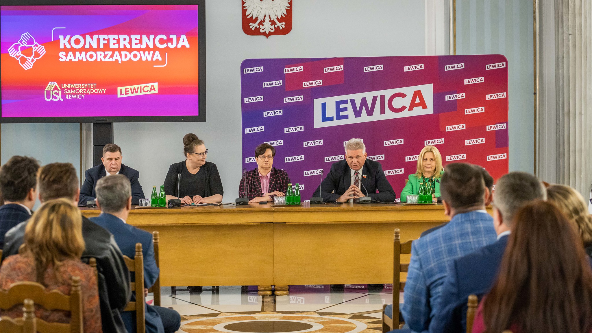 Panel dyskusyjny podczas konferencji samorządowej Lewicy; pięć osób siedzi za długim stołem, na tle baneru LEWICA i ekranu z napisem KONFERENCJA SAMORZĄDOWA.