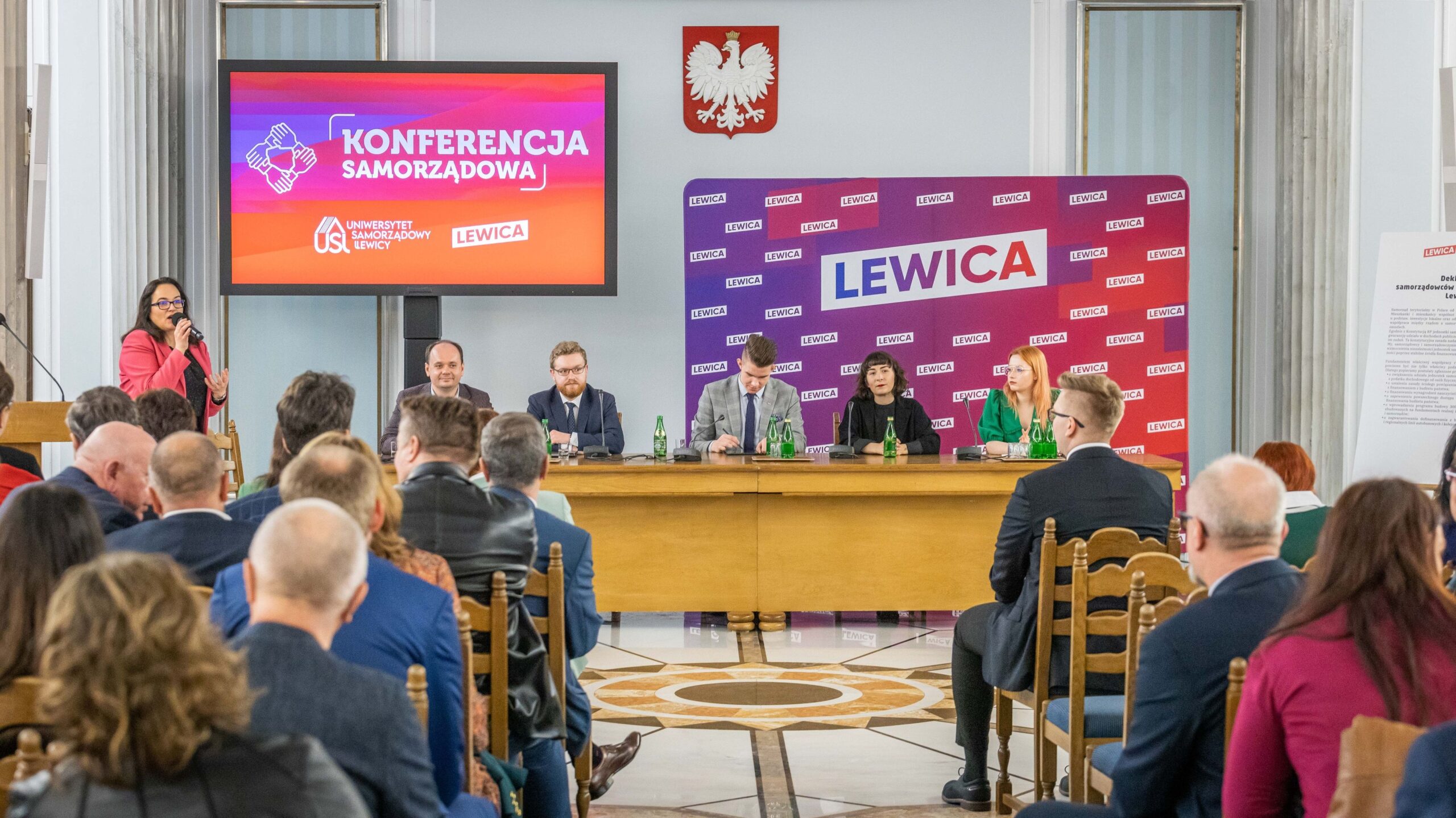 Panel konferencji samorządowej z prowadzącą kobietą z mikrofonem po lewej, pięcioma prelegentami przy stole i dużym ekranem z napisem „KONFERENCJA SAMORZĄDOWA”, w tle plakat Lewica i herb Polski.