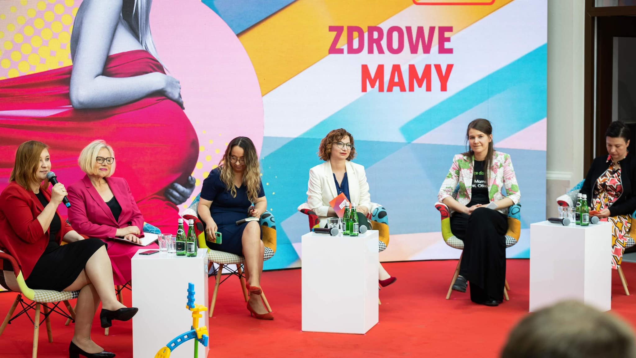 Panel dyskusyjny na konwencji "Mamy plan dla mam" – pięć kobiet siedzi na kolorowych krzesłach na czerwonym dywanie, a tło z napisem "ZDROWE MAMY" tworzy kolorową scenę.
