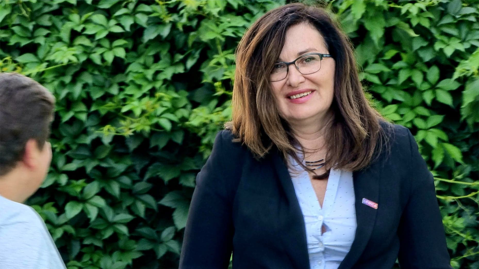 Na zdjęciu widoczna jest uśmiechnięta kobieta w eleganckim stroju, stojąca wśród zielonych liści.