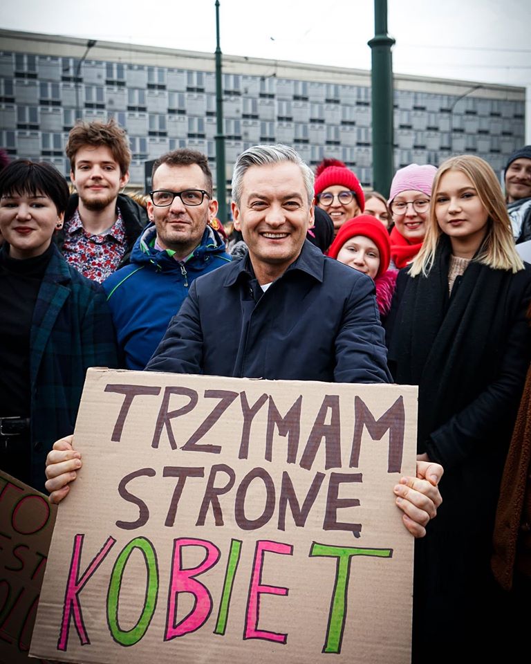 Grupa uczestników Manify „Feminizm dla klimatu, klimat na antykapitalizm” stoi na tle miasta, a mężczyzna w centrum trzyma karton z napisem „TRZYMAM STRONĘ KOBIET”.