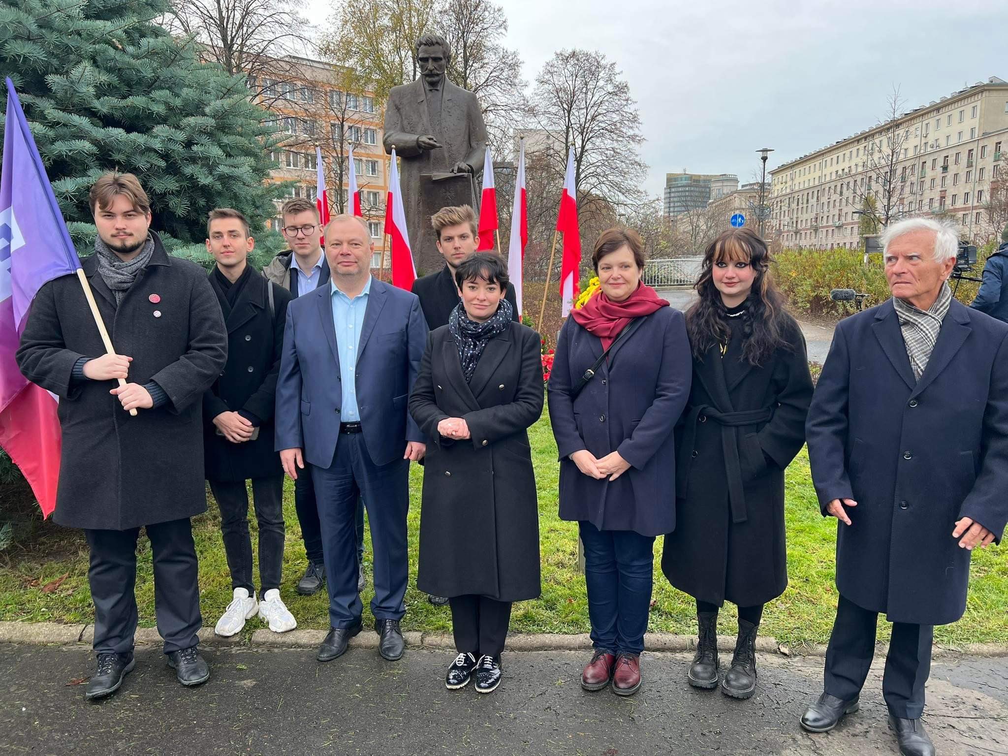 Na zdjęciu widoczna jest grupa ludzi stojących przed pomnikiem, z flagą w ręku i polskimi flagami w tle, z okazji jakiejś uroczystości.