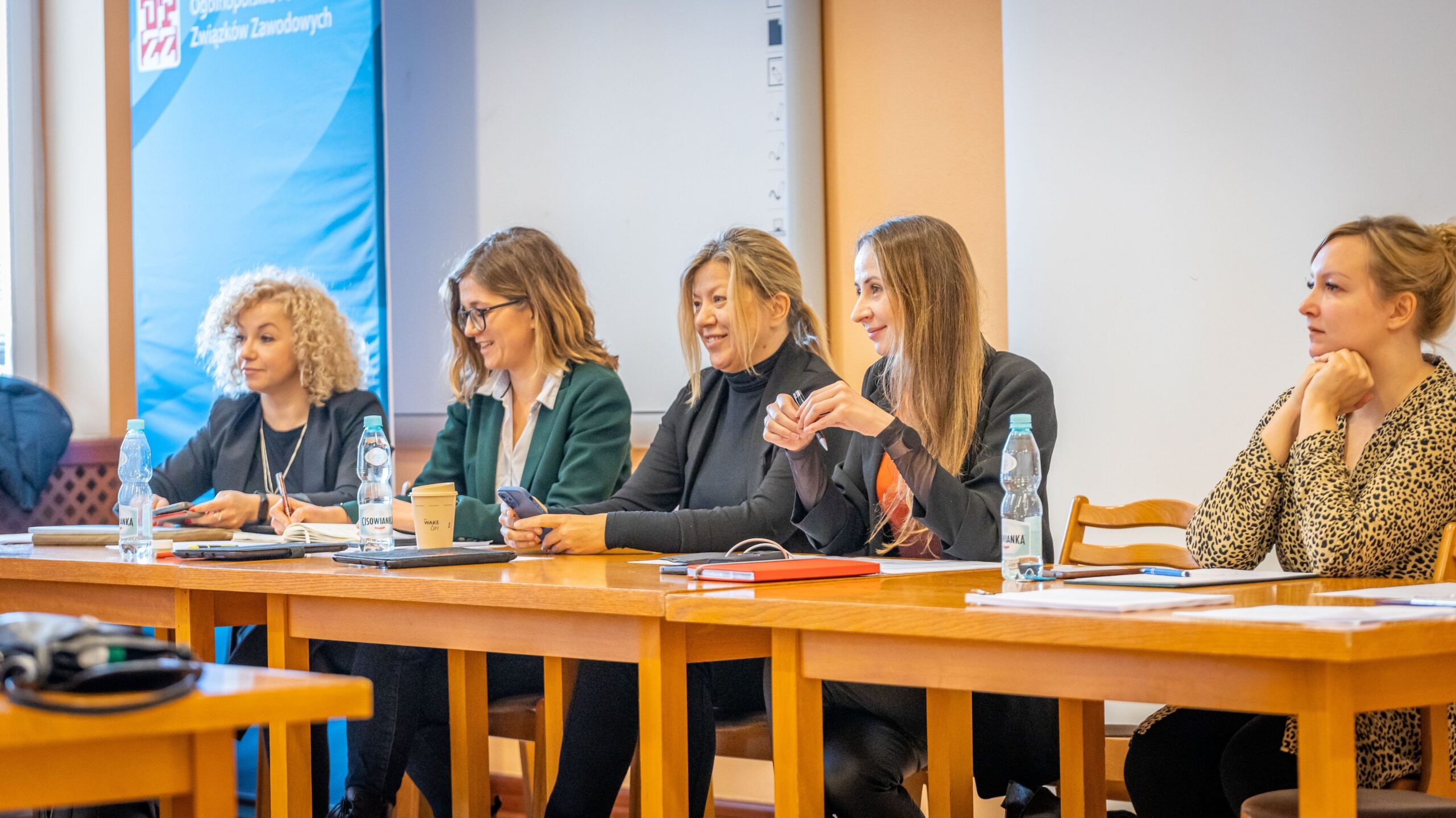 Kobiety siedzą przy długim stole podczas panelu o zmianach w Kodeksie pracy, z notatnikami, telefonami i butelkami wody, a w tle widać błękitny baner z logo związków zawodowych.
