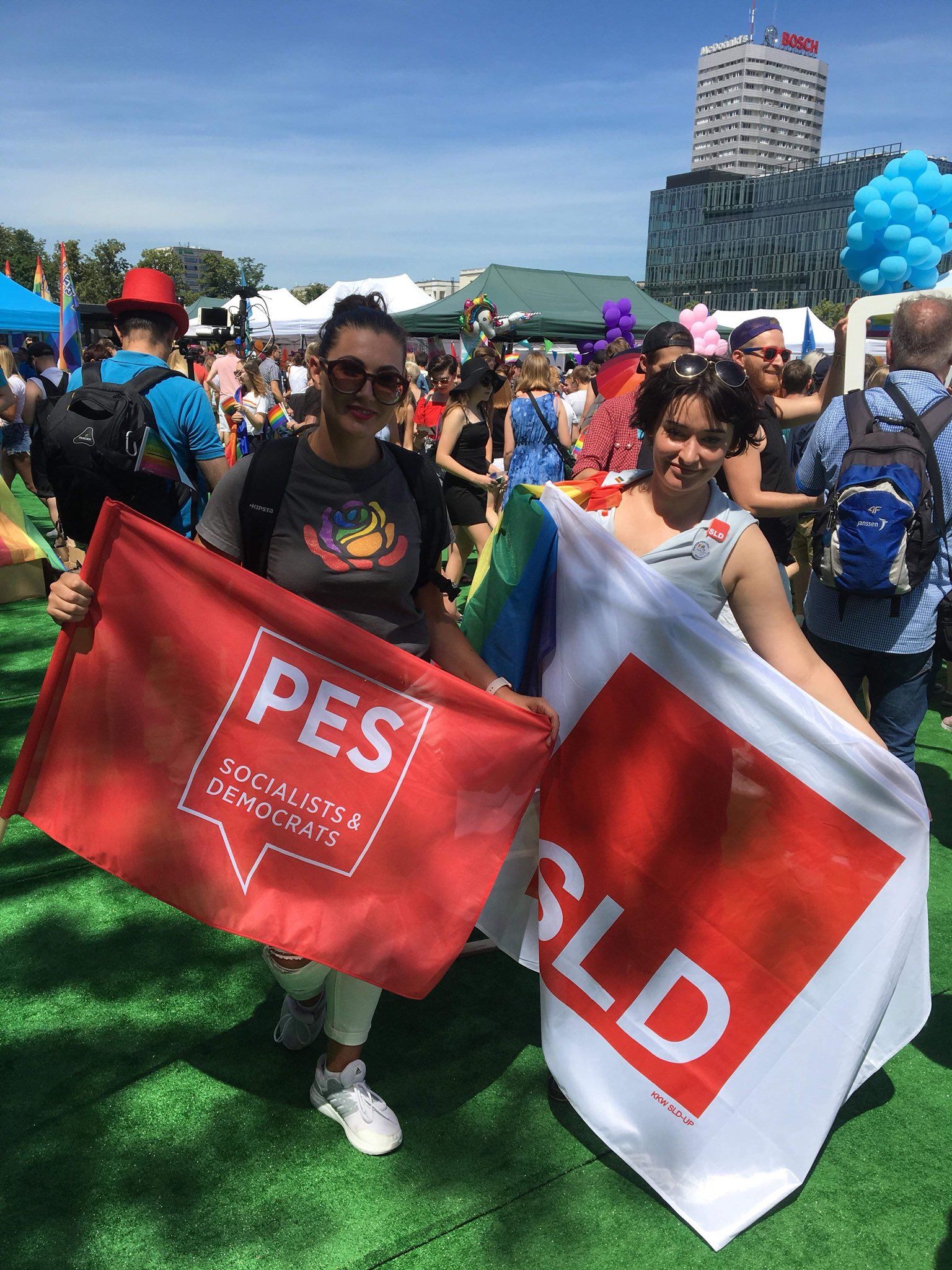 Dwie kobiety stoją na zielonej nawierzchni podczas 18. Parady Równości, trzymając flagi PES (Socialists & Democrats) i SLD wśród tłumu z namiotami i balonami.