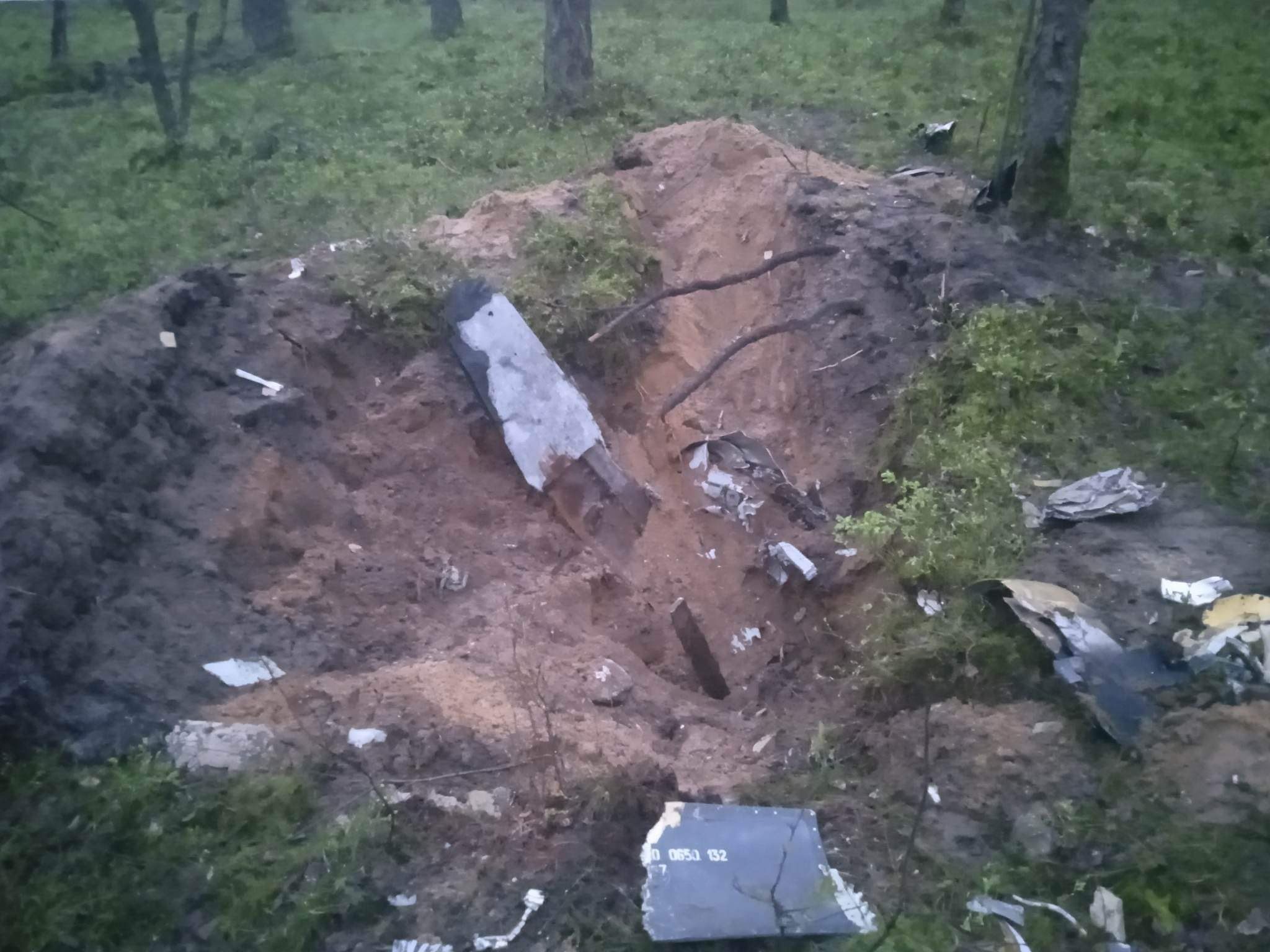 Zdjęcie przedstawia krater w lesie, otoczony pozostałościami metalowych fragmentów, które mogą pochodzić z opadłego obiektu.