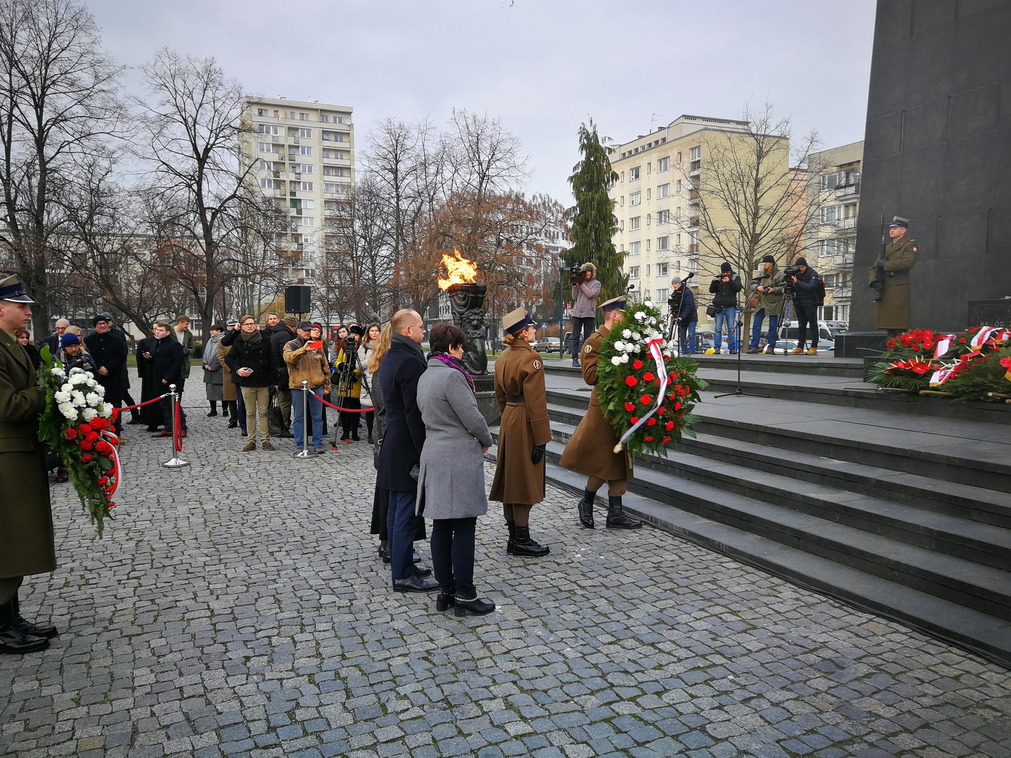 Uroczystość upamiętniająca 75. rocznicę wyzwolenia obozu Auschwitz-Birkenau, podczas której osoby w mundurach i cywile składają wieńce przed pomnikiem z płomieniem, na tle schodów i zgromadzonych mediów.