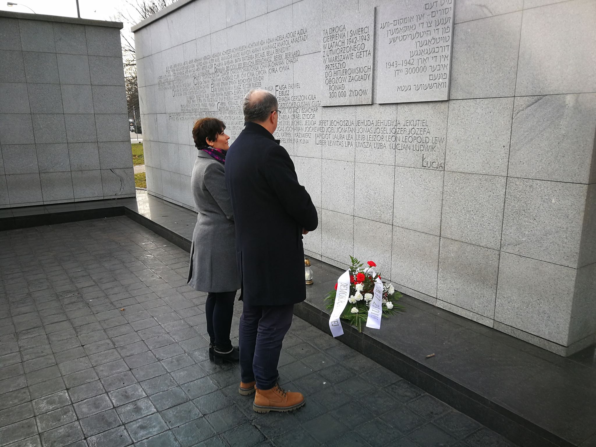 Para osób składa kwiaty przed ścianą memorialną z tablicami z nazwiskami upamiętniającymi ofiary Auschwitz-Birkenau.