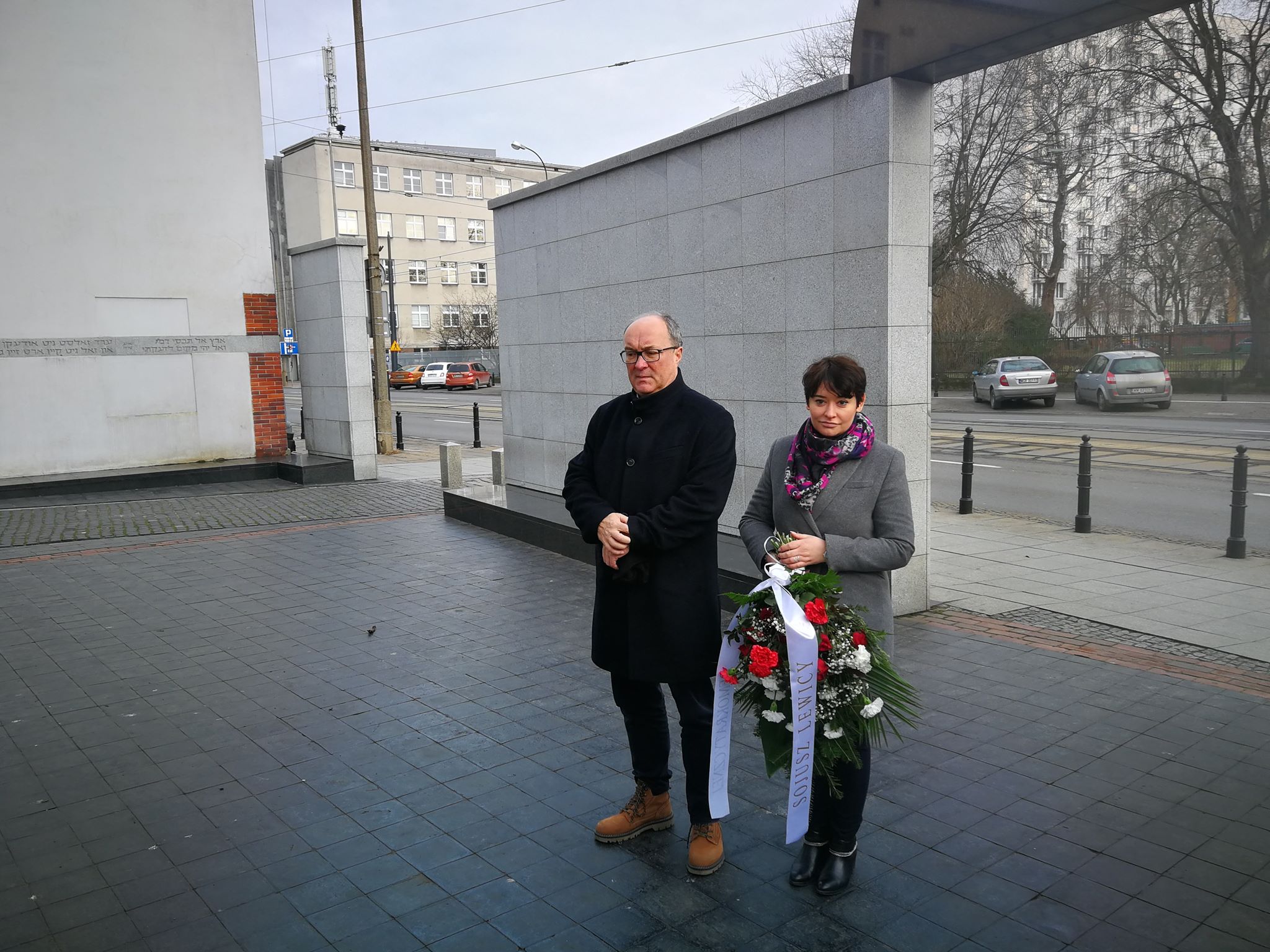 Dwie osoby stoją przed memorialną ścianą, trzymając wieniec z czerwonych i białych kwiatów podczas obchodów 75. rocznicy wyzwolenia obozu Auschwitz-Birkenau.