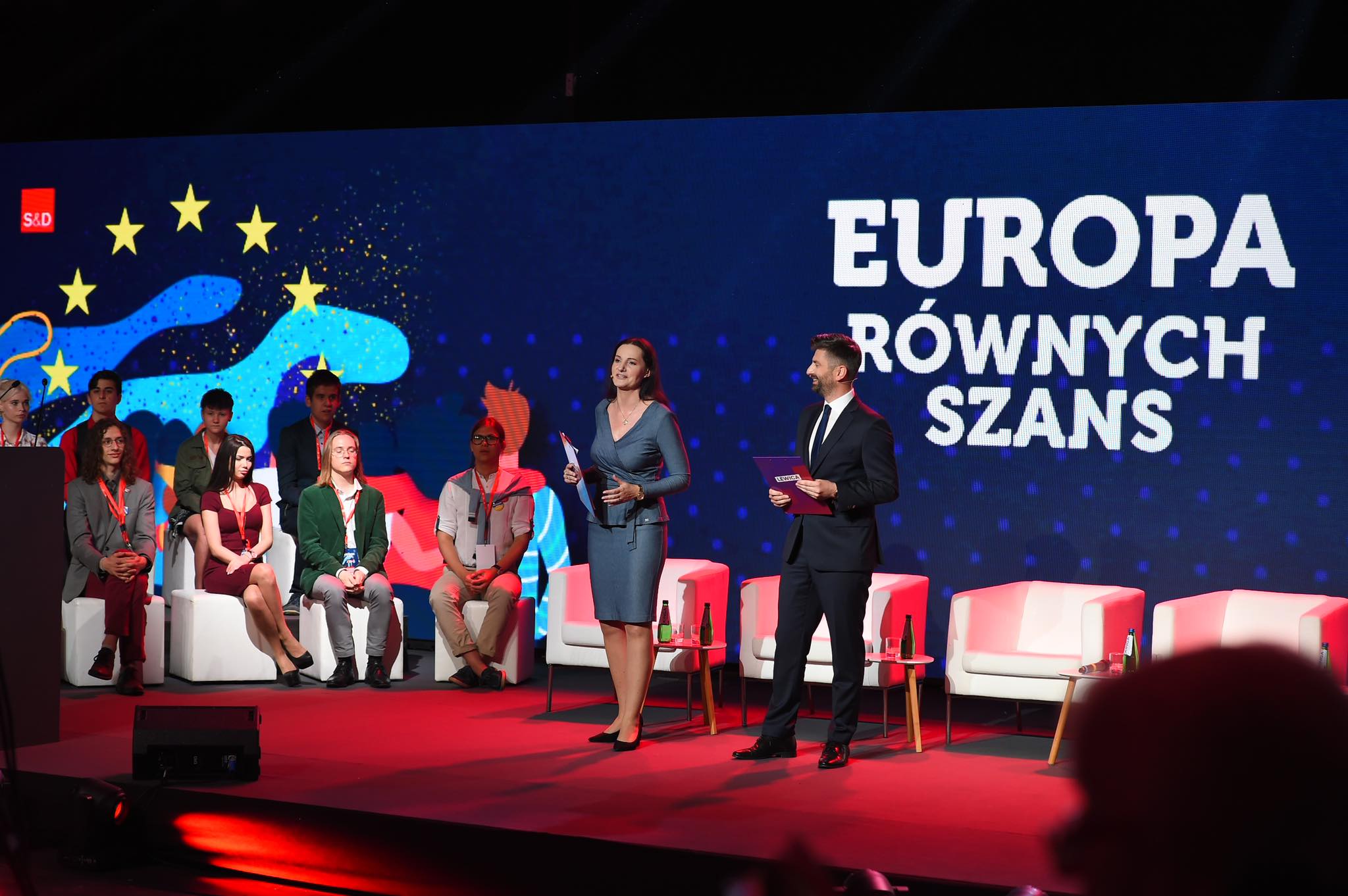 Na zdjęciu widoczni są prowadzący wydarzenie, mówiący do publiczności, a w tle znajduje się duży ekran z napisem