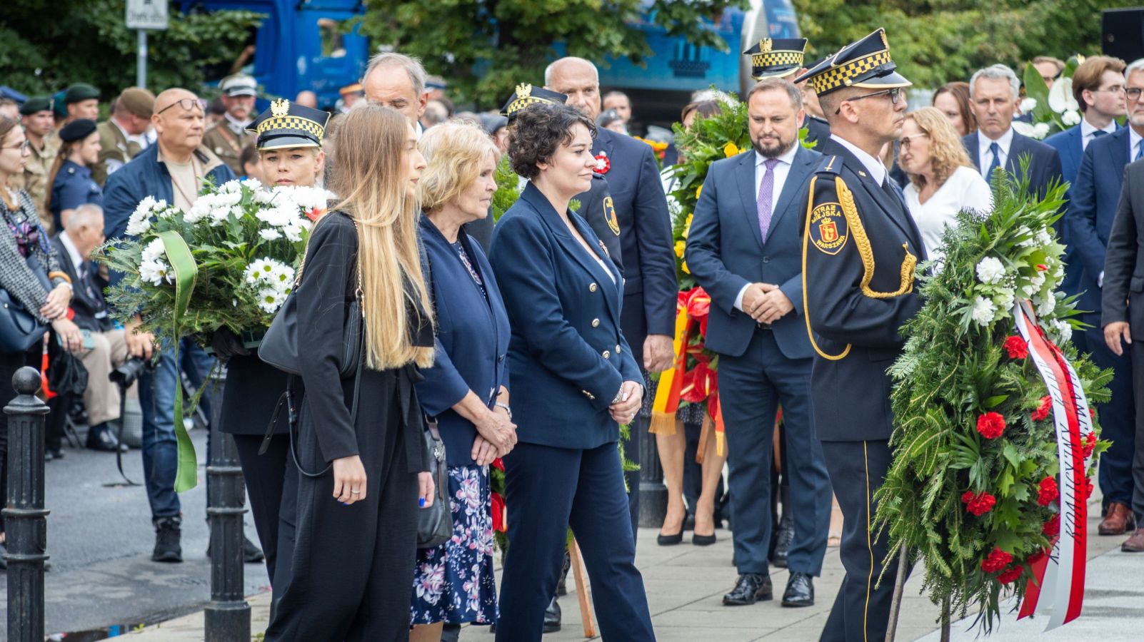 Uczestnicy obchodów 79. rocznicy Powstania Warszawskiego składają wieńce podczas uroczystości, stojąc wśród cywilów i funkcjonariuszy w mundurach.