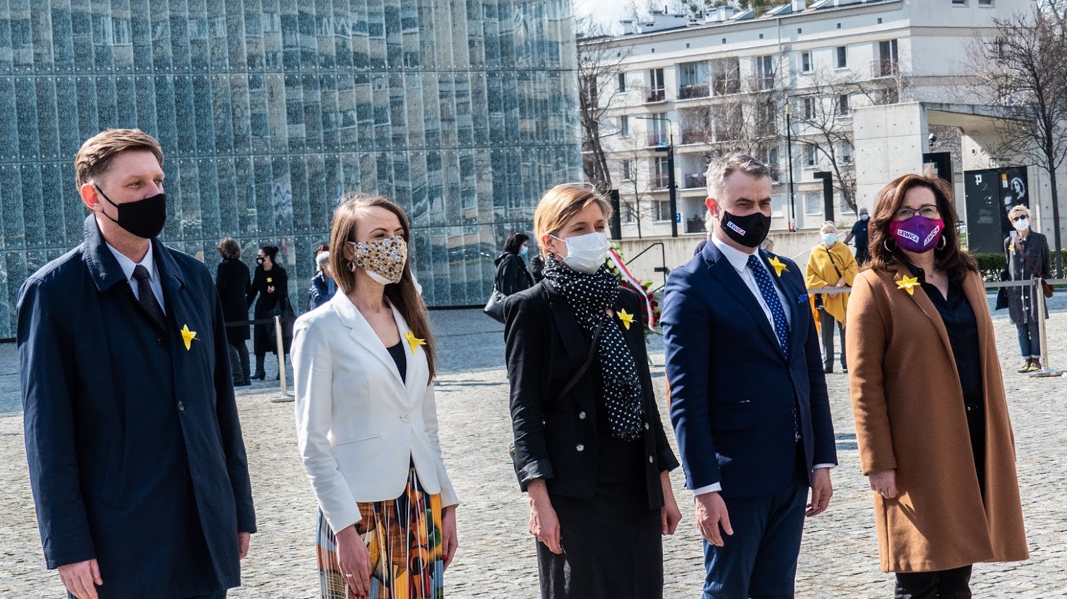 Grupa osób w maseczkach stoi na placu podczas obchodów 78. rocznicy Powstania w Getcie Warszawskim, z żółtymi przypinkami w kształcie gwiazd na ubraniach.
