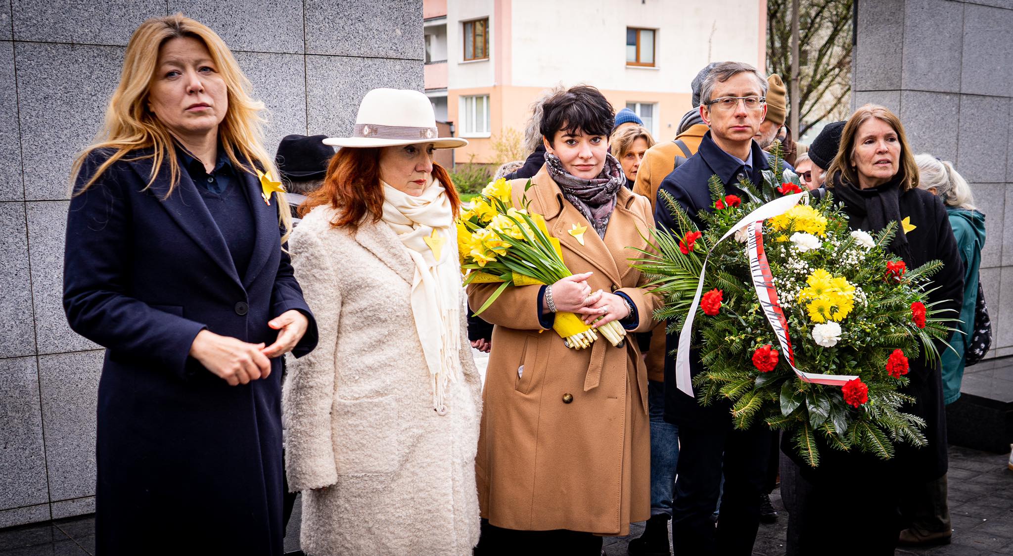 Grupa osób trzymająca żółte kwiaty oraz wieniec, ubrana w ciepłe ubrania i uczestnicząca w wydarzeniu pamięci.