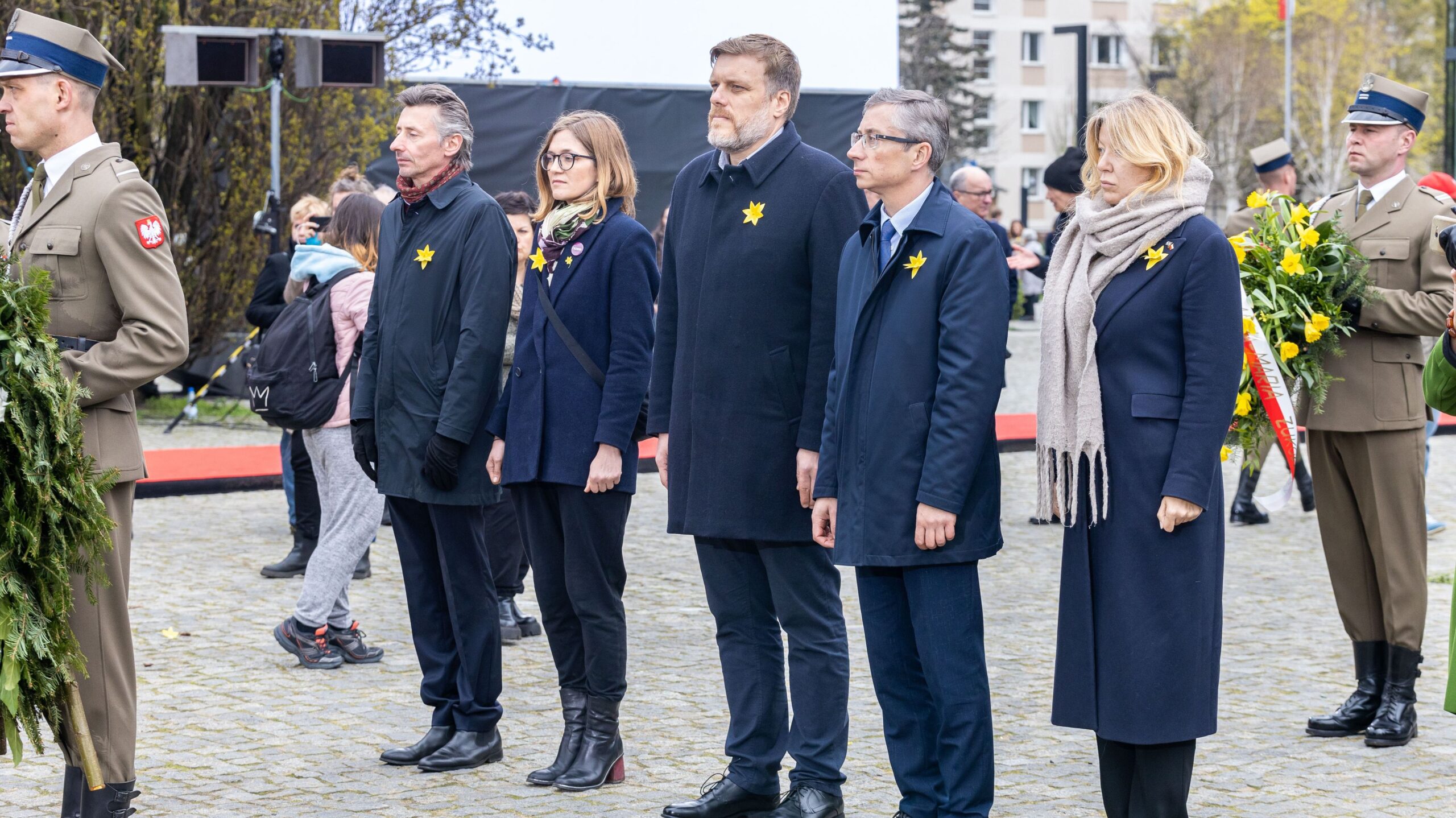 Na zdjęciu widoczni są uczestnicy uroczystości, stojący w kolejce przed trybuną, z kwiatami i emblematami w żółtym kolorze.