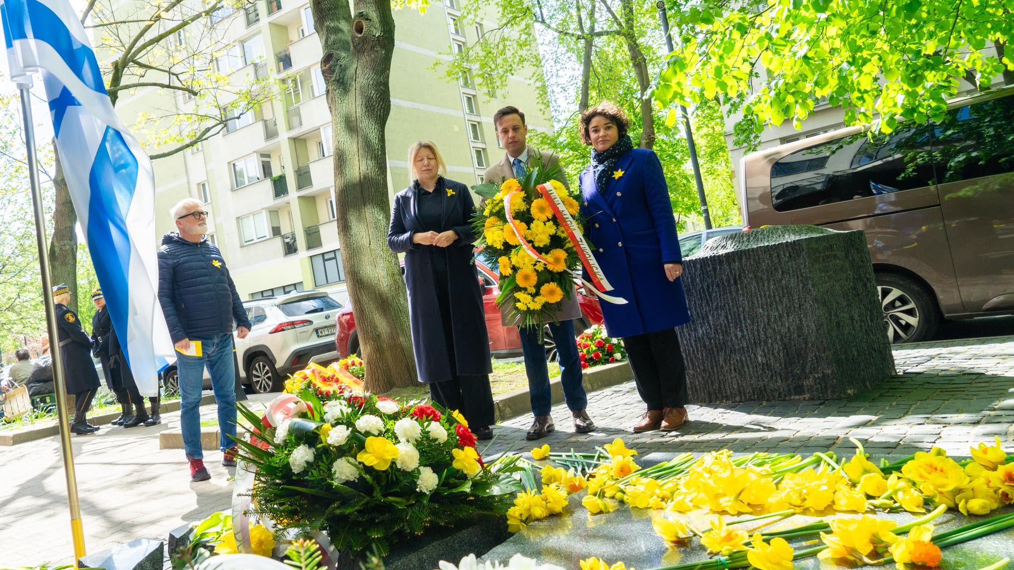 Grupa osób składa kwiaty i wieńce podczas obchodów 81. rocznicy powstania w getcie warszawskim, przy fladze i w otoczeniu zieleni.