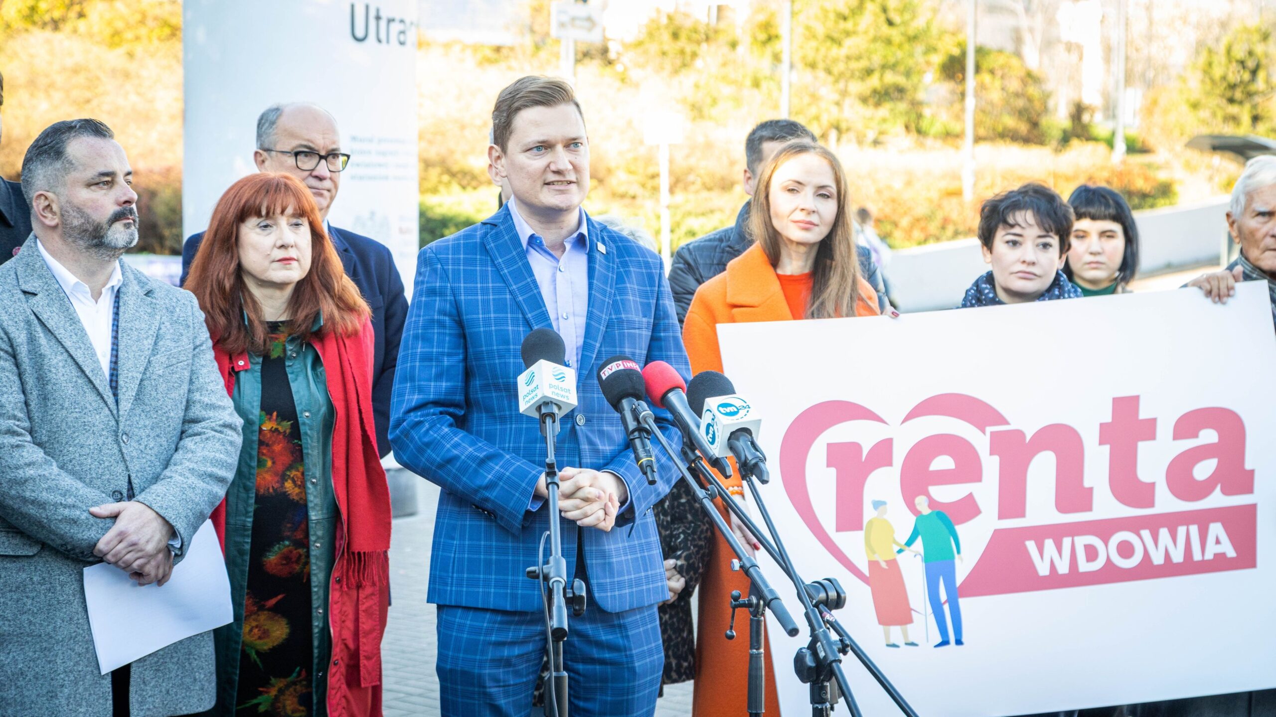 Na zdjęciu widoczna jest grupa ludzi, w tym mężczyzna w niebieskim garniturze, który przemawia do zebranych, a za nim stoi transparent z napisem „renta wdowia”.