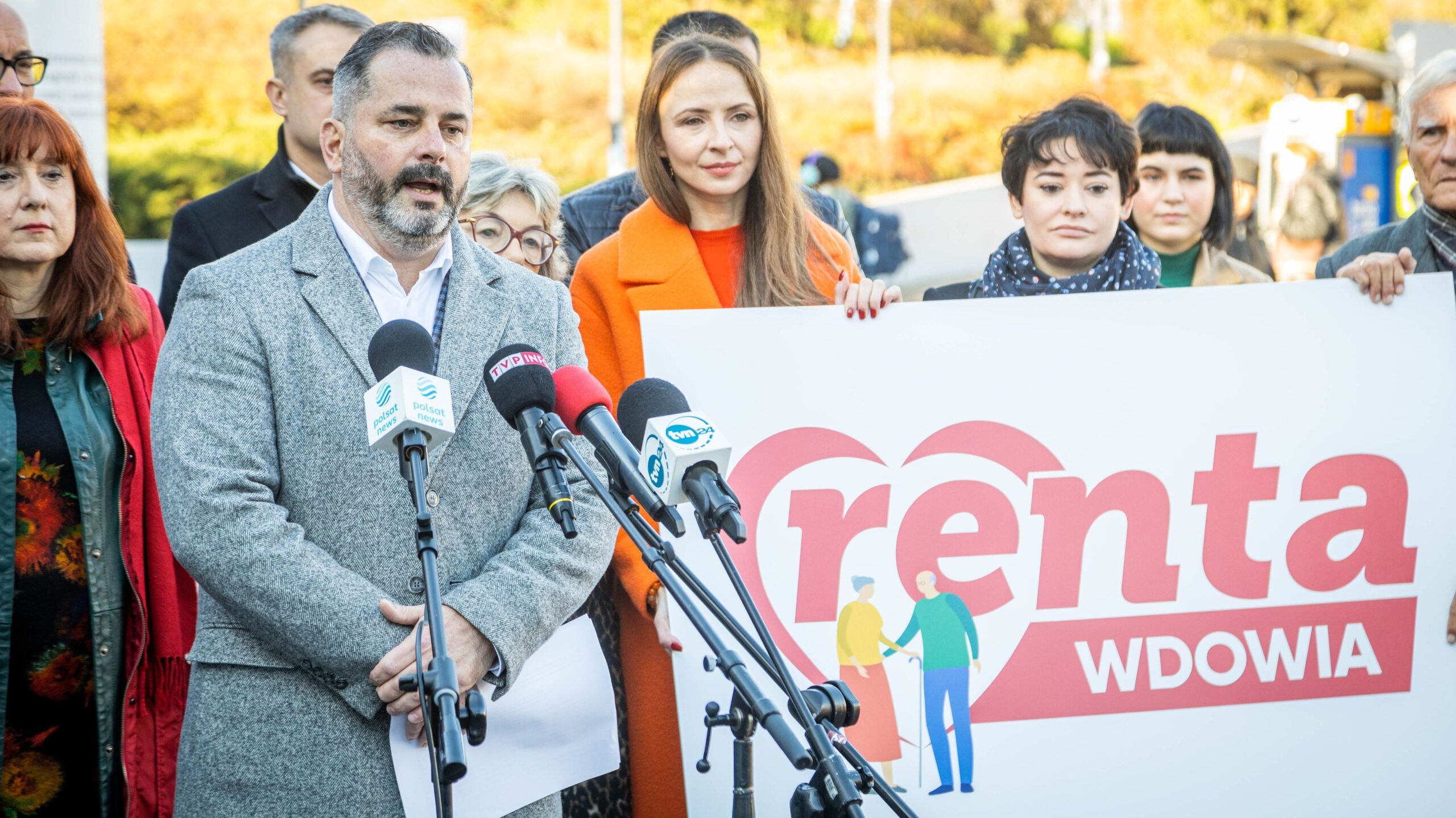 Konferencja prasowa dotycząca renty wdowiej: mężczyzna w szarym płaszczu przemawia do mikrofonów, stojąc obok kobiet i dużego baneru z napisem „renta WDOWIA”.