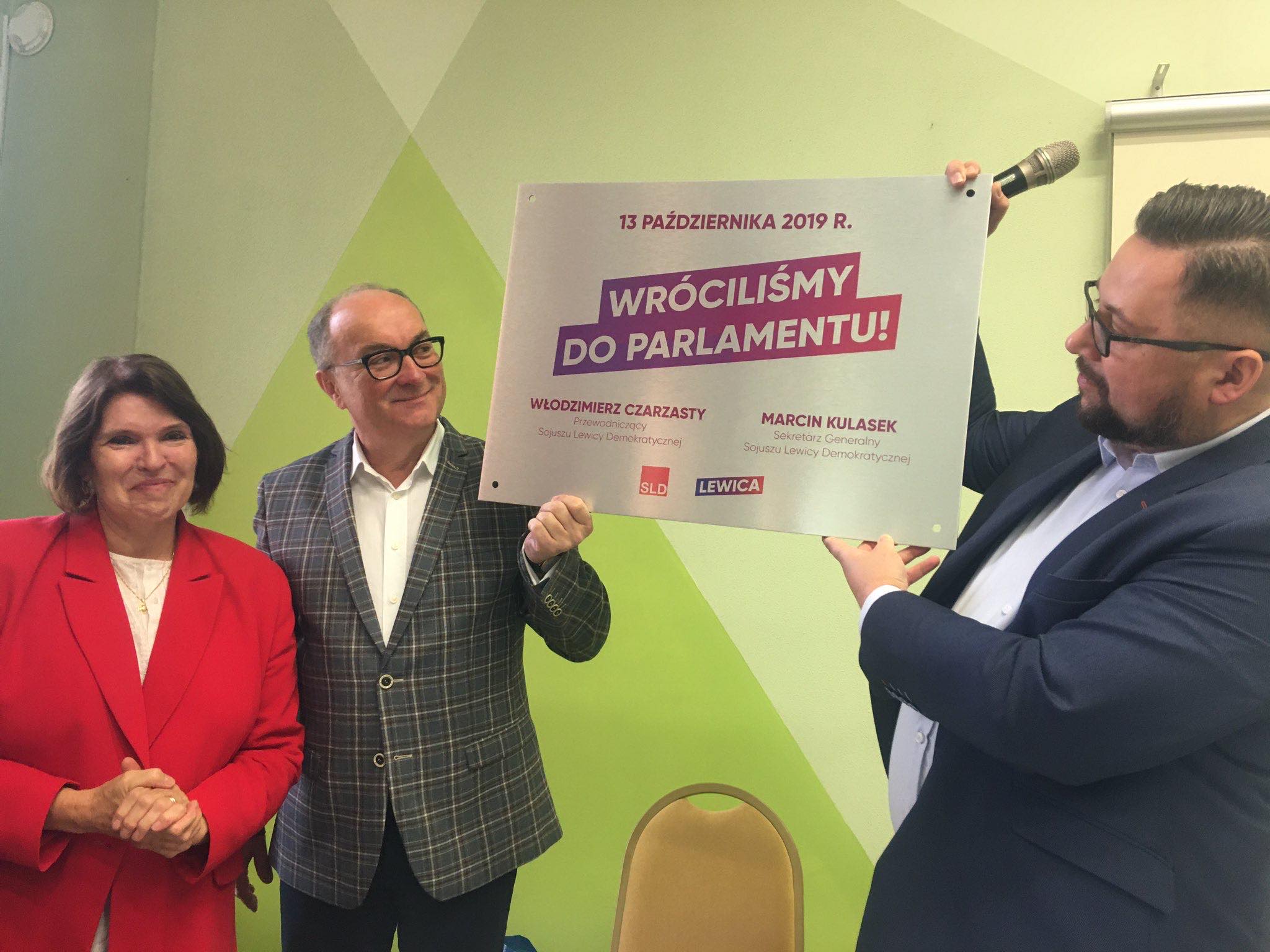 Trzy osoby stoją na zielonym tle i trzymają plakat z hasłem „Wróciliśmy do Parlamentu”.