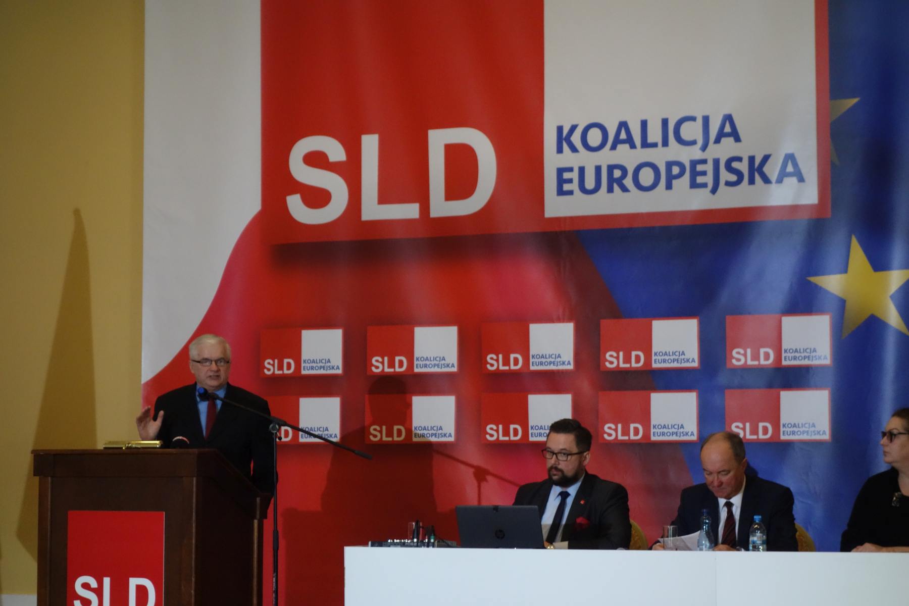 Kandydat SLD przemawia z mównicy podczas wydarzenia wyborczego do Parlamentu Europejskiego 2019, na tle dużych banerów z logotypem SLD i Koalicji Europejskiej, a przy stole siedzą dwie osoby z laptopem i dokumentami.