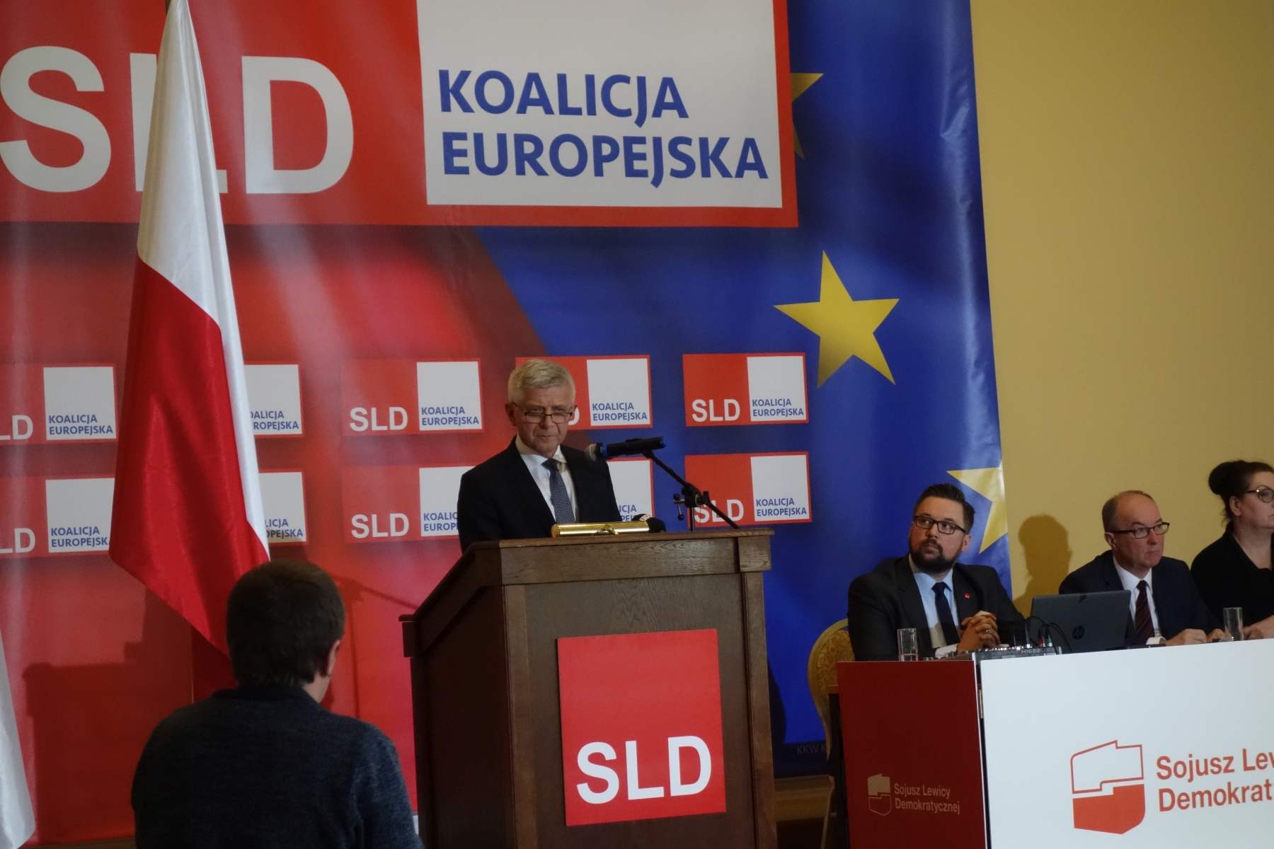 Mężczyzna w garniturze przemawia z mównicy podczas konferencji prasowej kandydatów SLD do Parlamentu Europejskiego w 2019 roku, na tle bannerów z logami SLD i Koalicji Europejskiej oraz flag Polski i Unii Europejskiej.