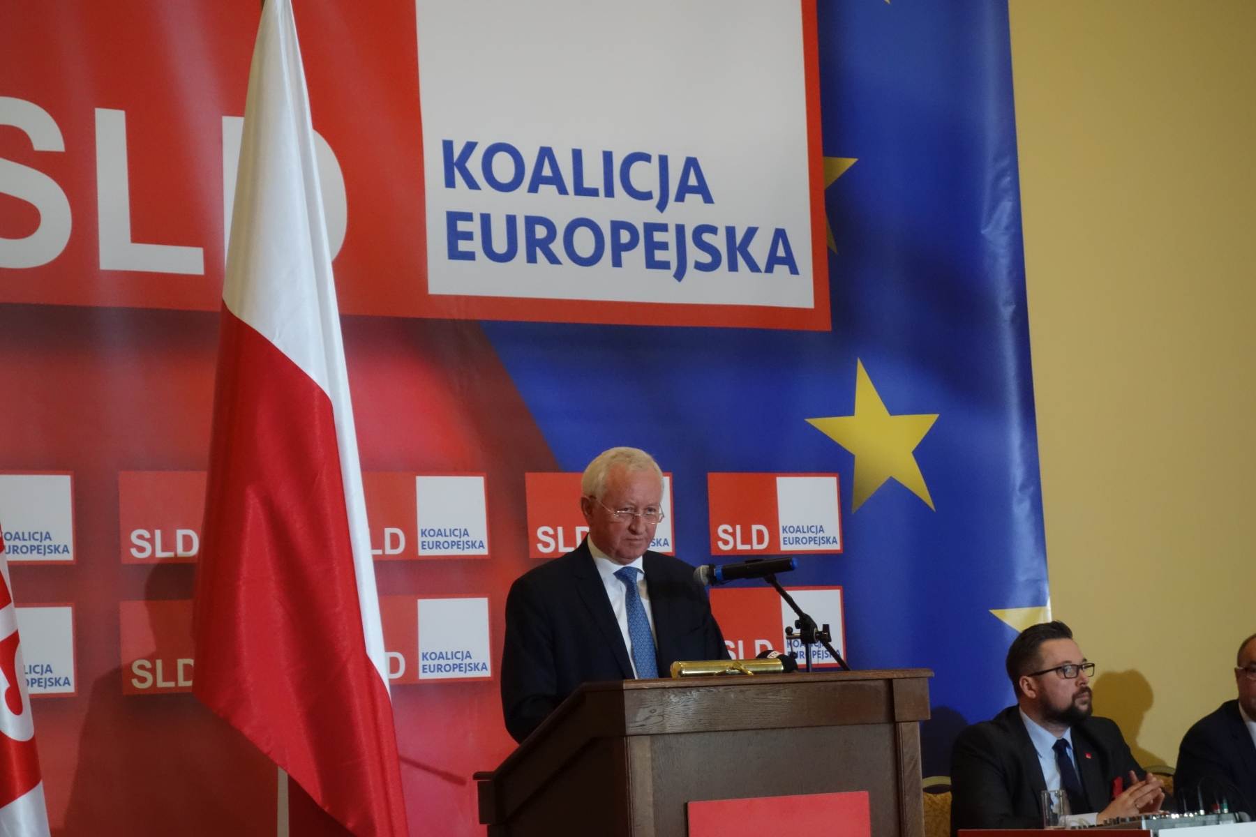 Kandydat SLD stojący przy mównicy podczas konferencji prasowej do wyborów do Parlamentu Europejskiego w 2019 roku, z flagą Polski i banerami Koalicji Europejskiej w tle.