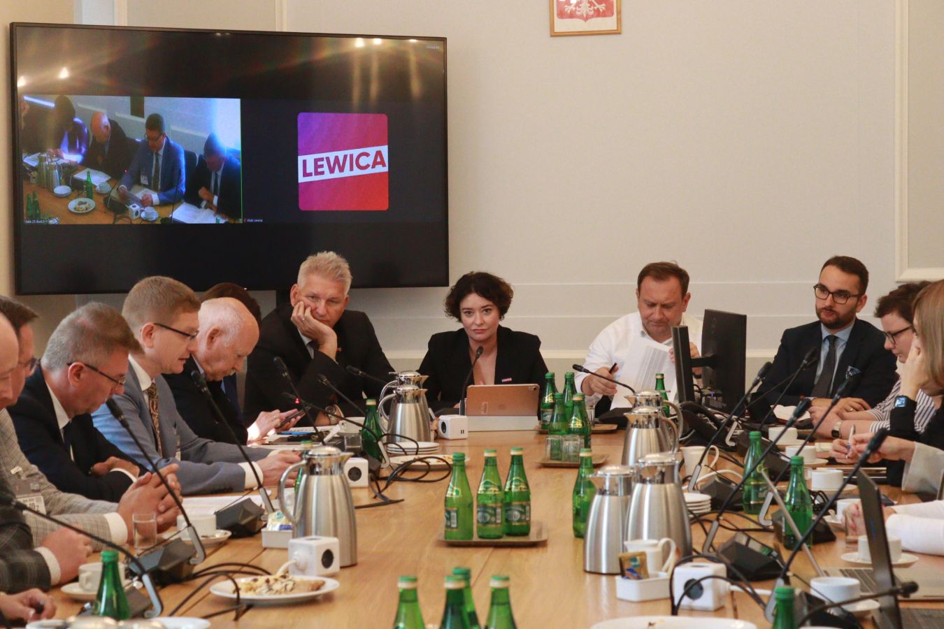 Grupa samorządowców siedzi przy długim stole w sali konferencyjnej podczas spotkania o finansowaniu samorządów; na dużym monitorze w tle wyświetlane jest posiedzenie i logo Lewica.