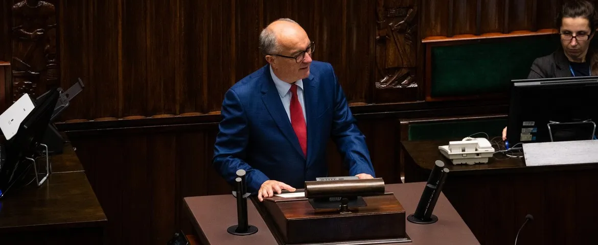 Mężczyzna w niebieskim garniturze i czerwonym krawacie stoi przy mównicy w sali z drewnianymi panelami i przemawia.
