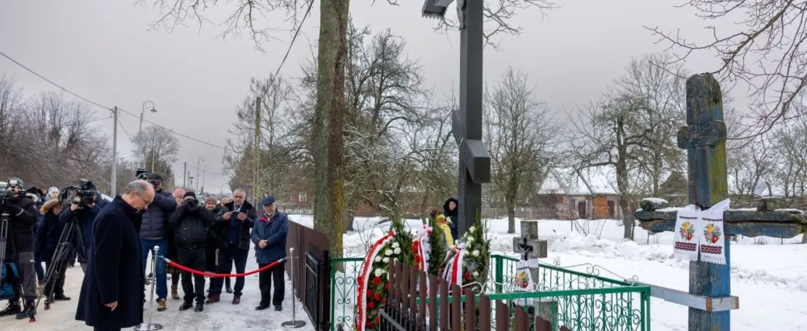 Grupa ludzi i dziennikarzy w zimowej scenerii składa wieńce biało-czerwone przy dużym czarnym krzyżu, stojąc za zielonym ogrodzeniem.