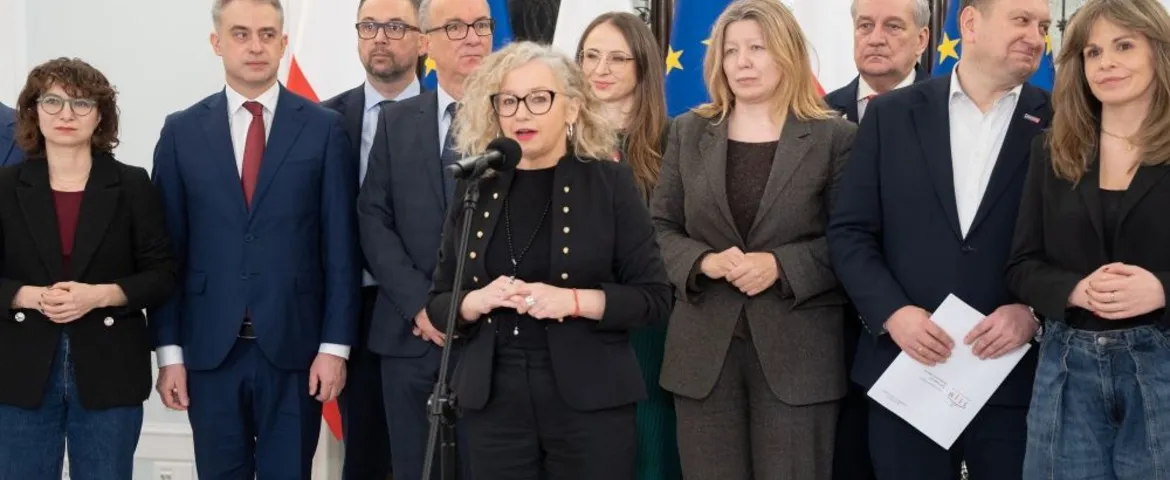 Grupa osób w formalnych ubraniach stoi na konferencji prasowej; na pierwszym planie blondynka z okularami przemawia do mikrofonu.