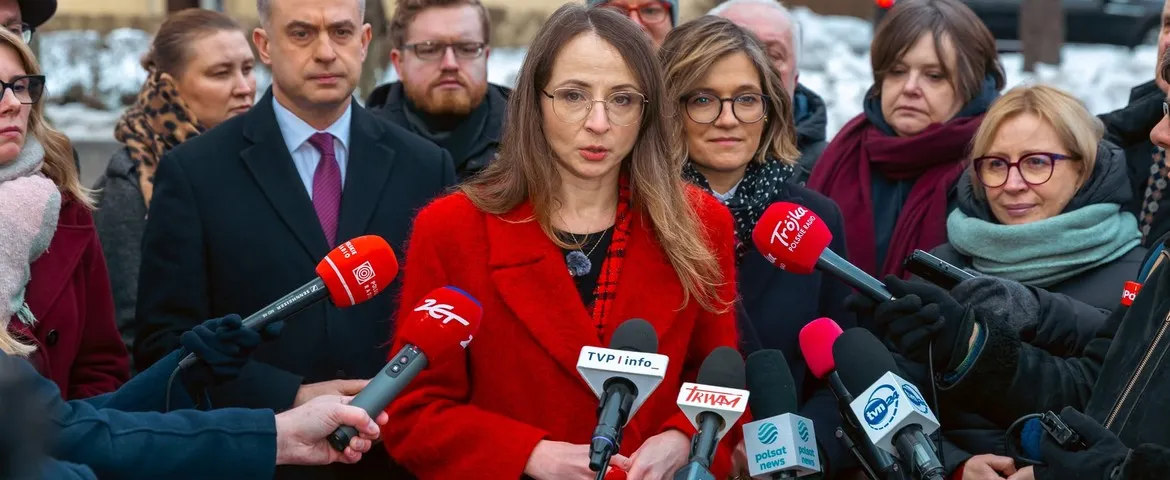 Kobieta w czerwonym płaszczu przemawia do zgromadzonych dziennikarzy, otoczona mikrofonami różnych stacji telewizyjnych.