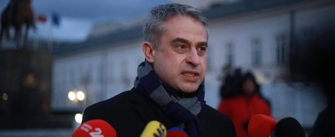 Mężczyzna w czarnym płaszczu i szaliku przemawia do kamery na tle budynku.