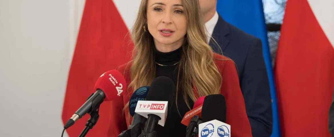 Kobieta w czerwonej marynarce przemawia podczas konferencji prasowej, stojąc przed mikrofonami z logotypami stacji telewizyjnych i flagami w tle.