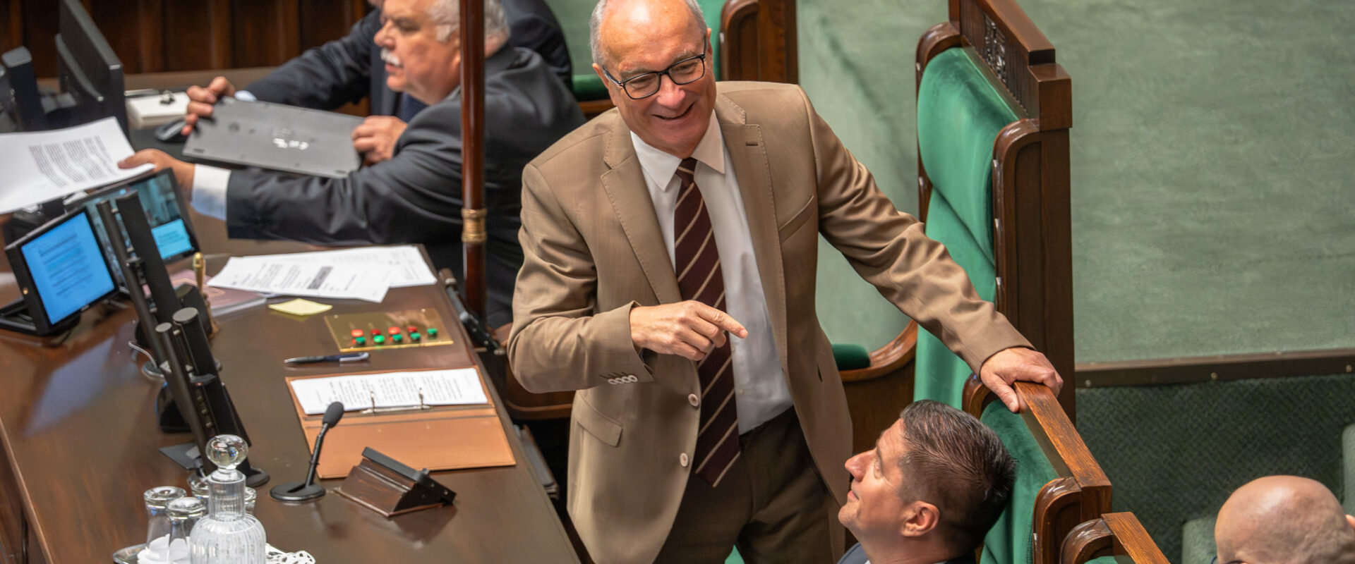 Mężczyzna w beżowym garniturze i czerwonym krawacie stoi przy biurku w sali plenarnej, uśmiechnięty, otoczony innymi politykami i dokumentami.