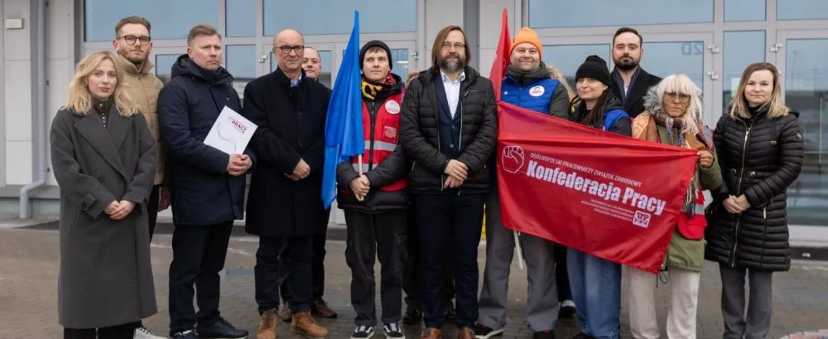 Grupa osób w zimowych ubraniach stoi przed wejściem do budynku, trzymając czerwony baner z napisem Konfederacja Pracy i niebieską flagę.