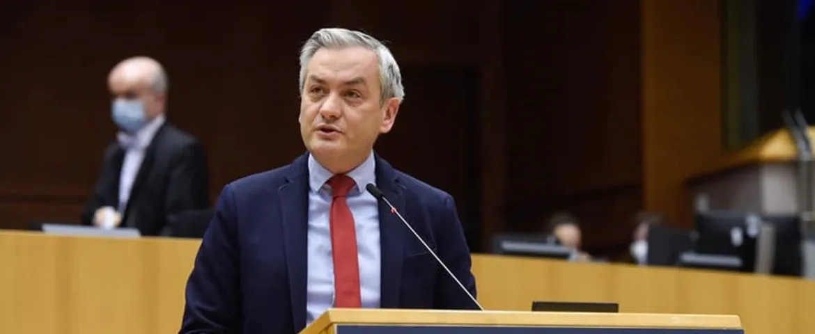 Mężczyzna w granatowym garniturze i czerwonym krawacie przemawia z mównicy podczas posiedzenia w sali obrad; w tle widoczni są inni uczestnicy.