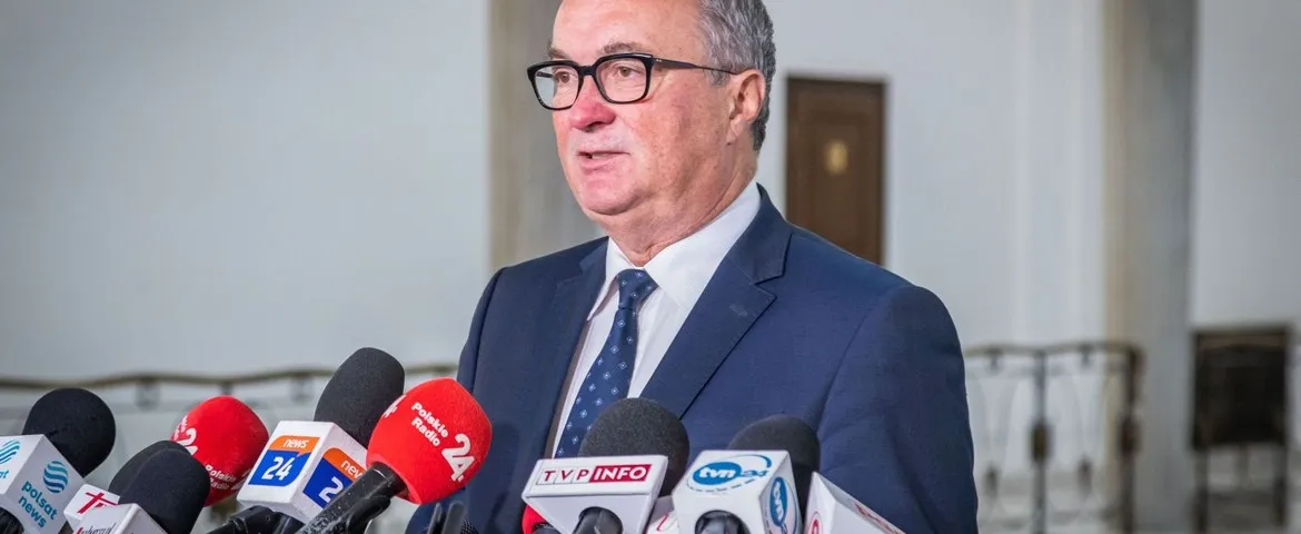 Mężczyzna w granatowym garniturze i okularach stoi przy zestawie mikrofonów podczas konferencji prasowej w jasnym wnętrzu.
