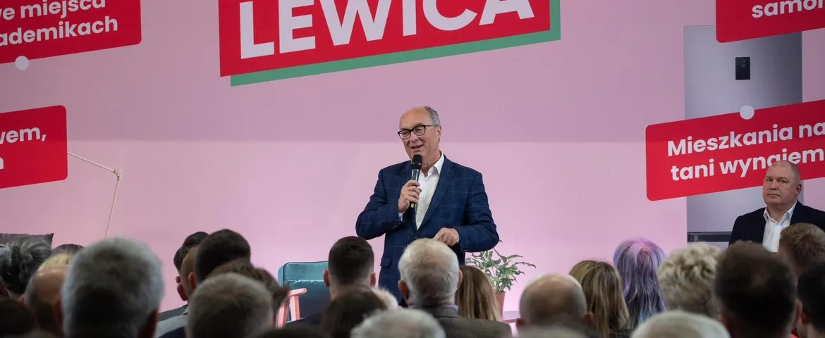Mężczyzna w granatowym garniturze przemawia do zgromadzonej publiczności na tle różowego scenicznego tła z dużym napisem Lewica.